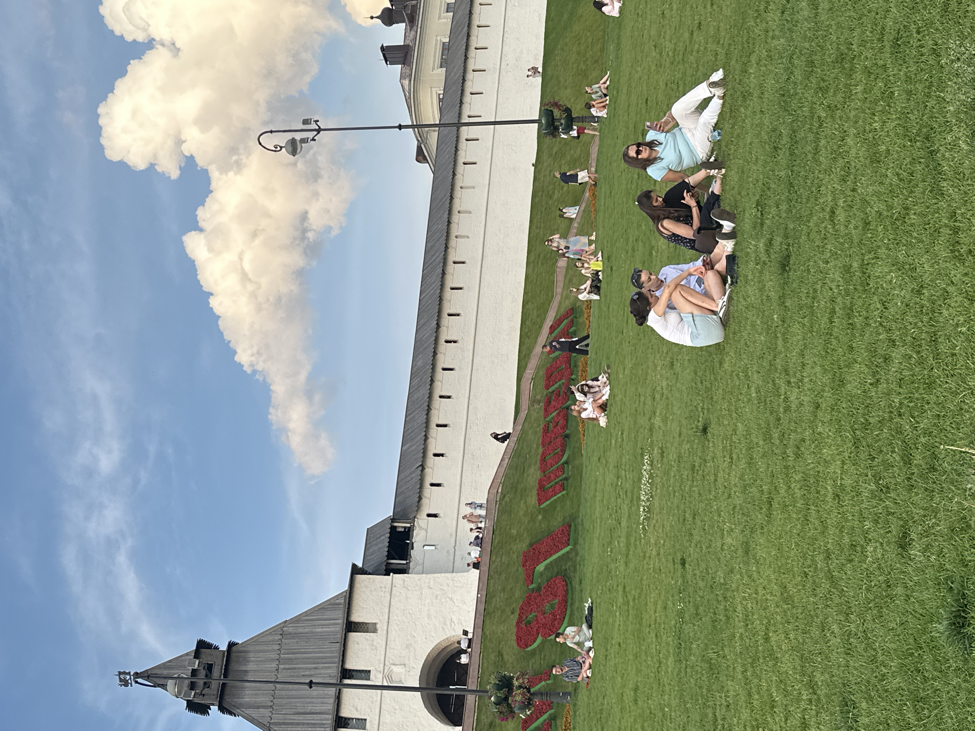 The image depicts a sunny day in Kazan, Russia, with a view of the Kazan Kremlin's white wall and a prominent wooden tower. In the foreground, a lush green lawn is dotted with people enjoying the outdoors. Several groups are seated on the grass, engaged in various activities. One group of four young women sits together, with one woman looking at her phone and another pointing. To the left, another group of three people sits, one of whom appears to be looking at a phone. Further back, scattered individuals and small groups are visible, some walking along a pathway or sitting on the grass. A large floral display in the shape of red numbers is visible on the grassy slope, partially obscured by people. The sky is a vibrant blue, with fluffy white clouds. The overall scene conveys a relaxed and pleasant atmosphere in a historical urban setting.