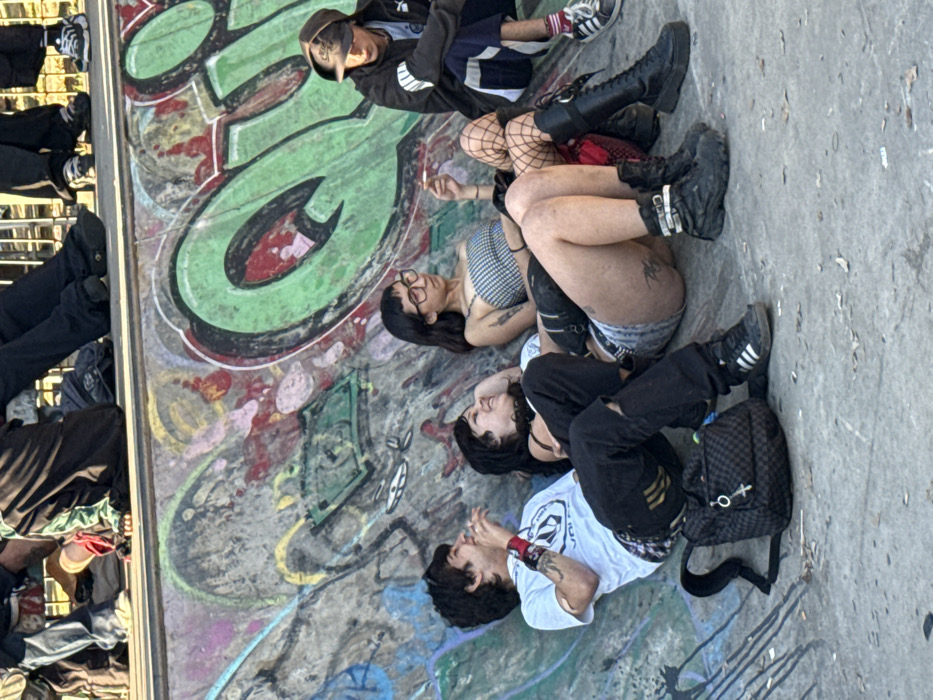 The image depicts four young adults resting within the Skatepark Parque Centenario in Buenos Aires, Argentina. The background consists of a curved concrete skate park wall heavily adorned with colorful graffiti tags and designs, including large green bubble letters and a drawing resembling a US dollar bill. The foreground shows a light-colored concrete surface.
On the far left, a male subject with dark hair leans against the graffiti-covered wall, wearing a white t-shirt with a black "Volcom" logo, ripped black jeans, and black Adidas sneakers. Tattoos are visible on his right arm and wrist. His right hand is near his mouth. A black and white checkered backpack with a metal charm hangs on the ground beside him.
Next to him, a female subject with dark hair leans back against the wall, smiling and looking towards the male on the left. She wears a dark top and dark pants.
To her right, a second female subject sits upright, wearing glasses, a black and white gingham crop top, light-colored denim shorts, and black combat boots. Her left leg is partially covered by a black fishnet stocking, and multiple tattoos are visible on her legs and arms. She holds a lit cigarette in her right hand. A black fanny pack is slung across her chest.
On the far right, a male subject wearing a dark hooded jacket, dark pants with white stripes, and dark sneakers sits with a baseball cap worn backwards. He leans forward, looking downwards.
Above the resting individuals, blurred legs of other people are visible sitting along the top edge of the skate park wall. The scene is illuminated by bright daylight.