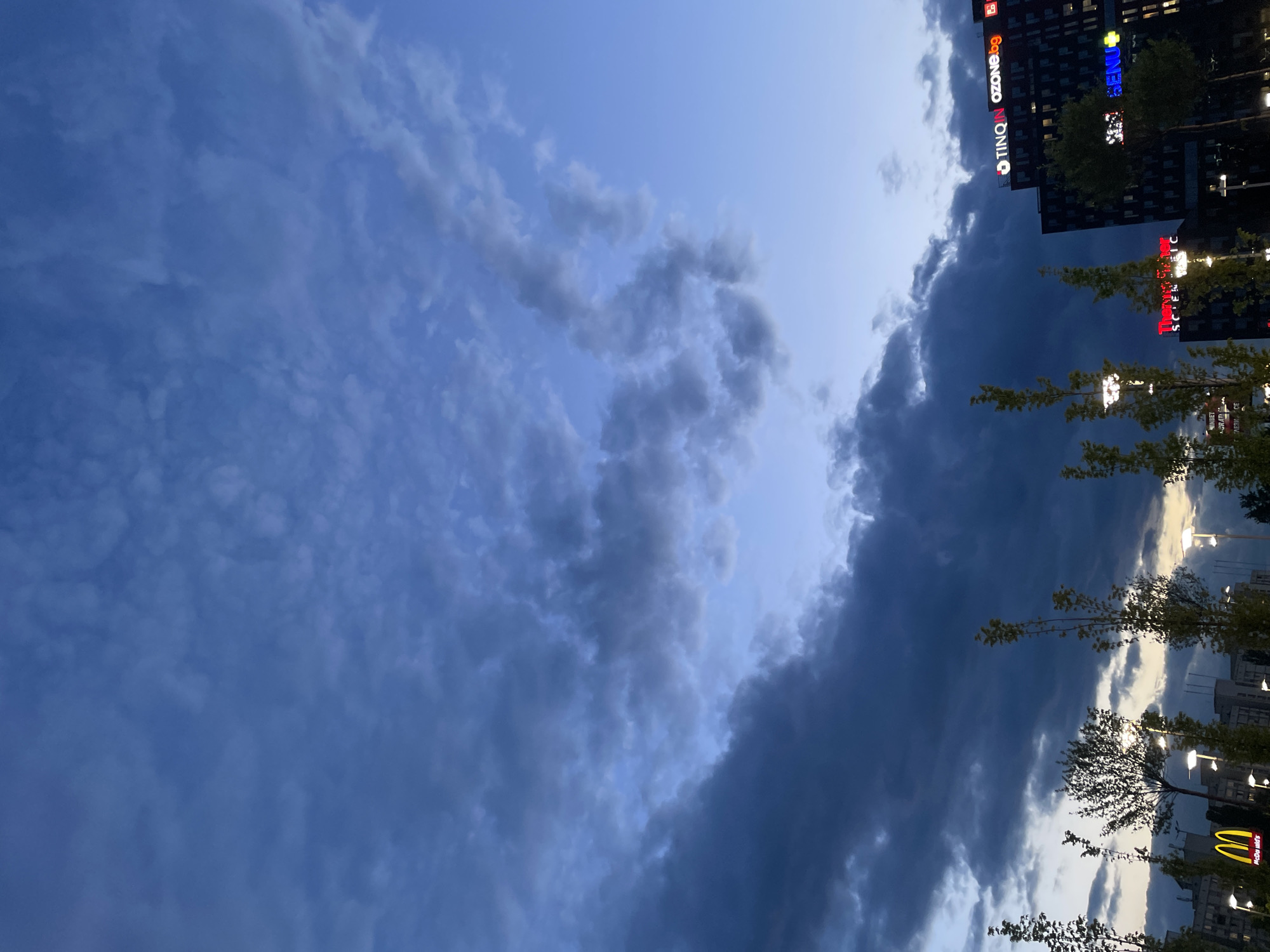 The image captures a partially cloudy sky above a city environment. Buildings with illuminated signs, including "TINQ" "ozone.og" and "SENU+" are visible. There are also trees with yellowing leaves. A McDonalds sign is partially visible in the lower right corner. The sky is a mix of blue and dark grey clouds, suggesting a change in weather or the end of the day. The photo appears to be taken from a ground-level perspective, looking upwards. The location is Unknown, Unknown.