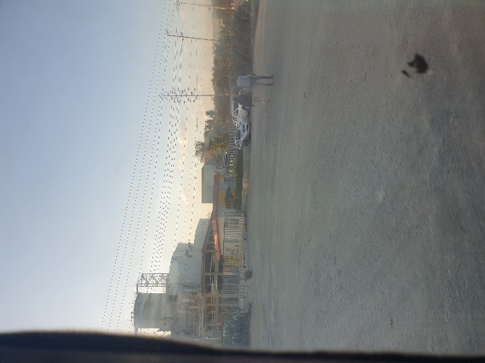 A sideways landscape shows a construction site with large cylindrical tanks and scaffolding under a pale sky with power lines. A road with a dark spot dominates the foreground. A person walks near parked cars in the middle ground, and low vegetation lines the back.