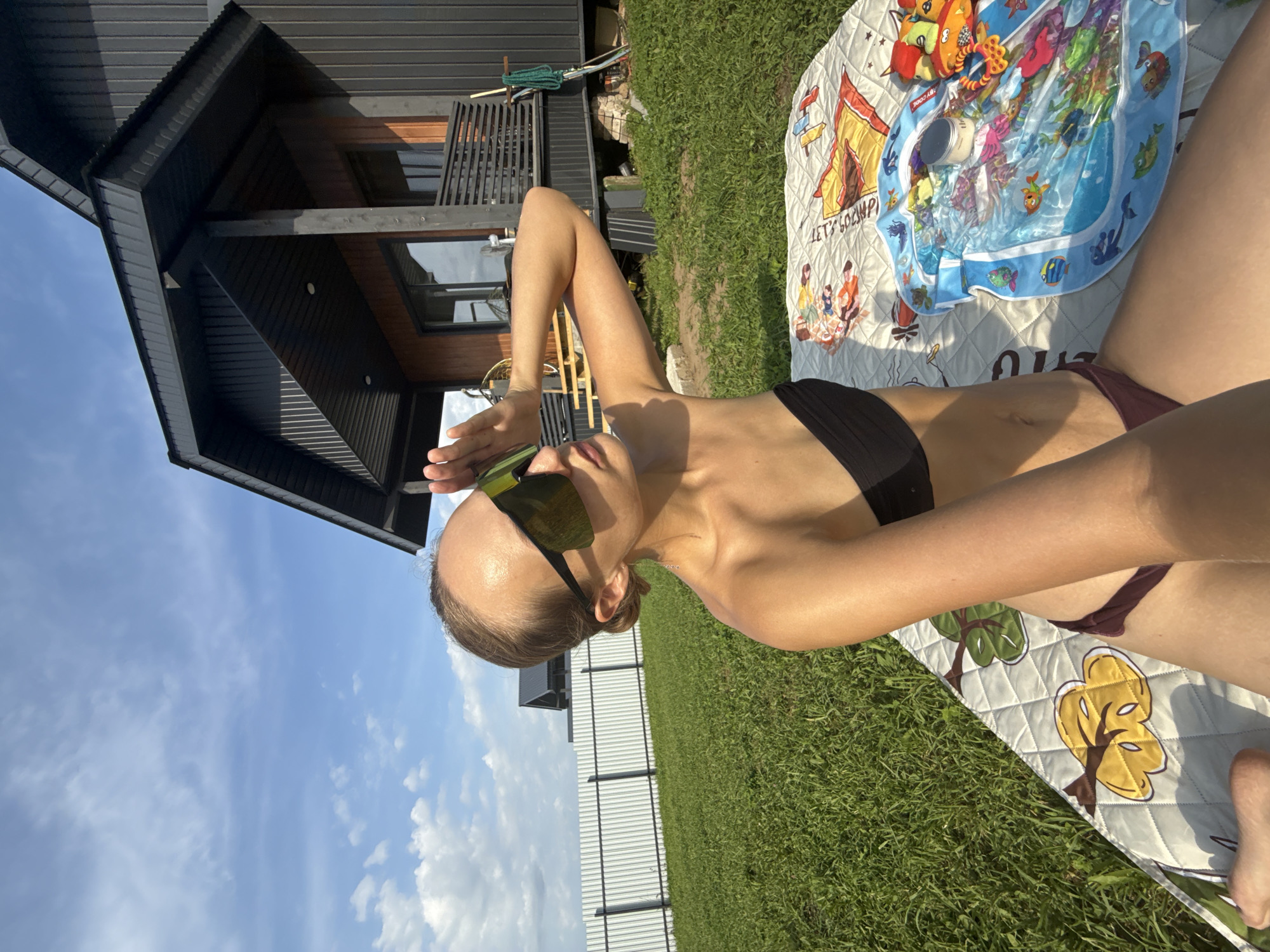 A young woman in a black bikini and large sunglasses is sitting on a blanket on a grassy lawn in front of a modern wooden house. She is shielding her eyes from the sun with her right hand. The blanket has a quilted pattern with colorful cartoonish images of fruits and possibly people, along with a small inflatable pool with fish depicted on it. A white fence runs along the edge of the lawn. The sky is blue with scattered clouds. The location is identified as Podshivalovo, Russia.