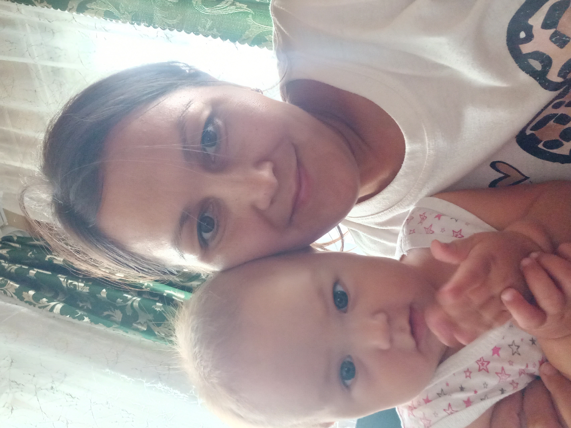 A selfie shows a woman and a baby. The woman, with brown hair and dark eyes, is smiling. She is wearing a white t-shirt. The baby, with blonde hair and blue eyes, is looking at the camera. The baby is wearing a white onesie with pink stars. They are indoors, with curtains in the background. The location is identified as Идрисово, Russia.