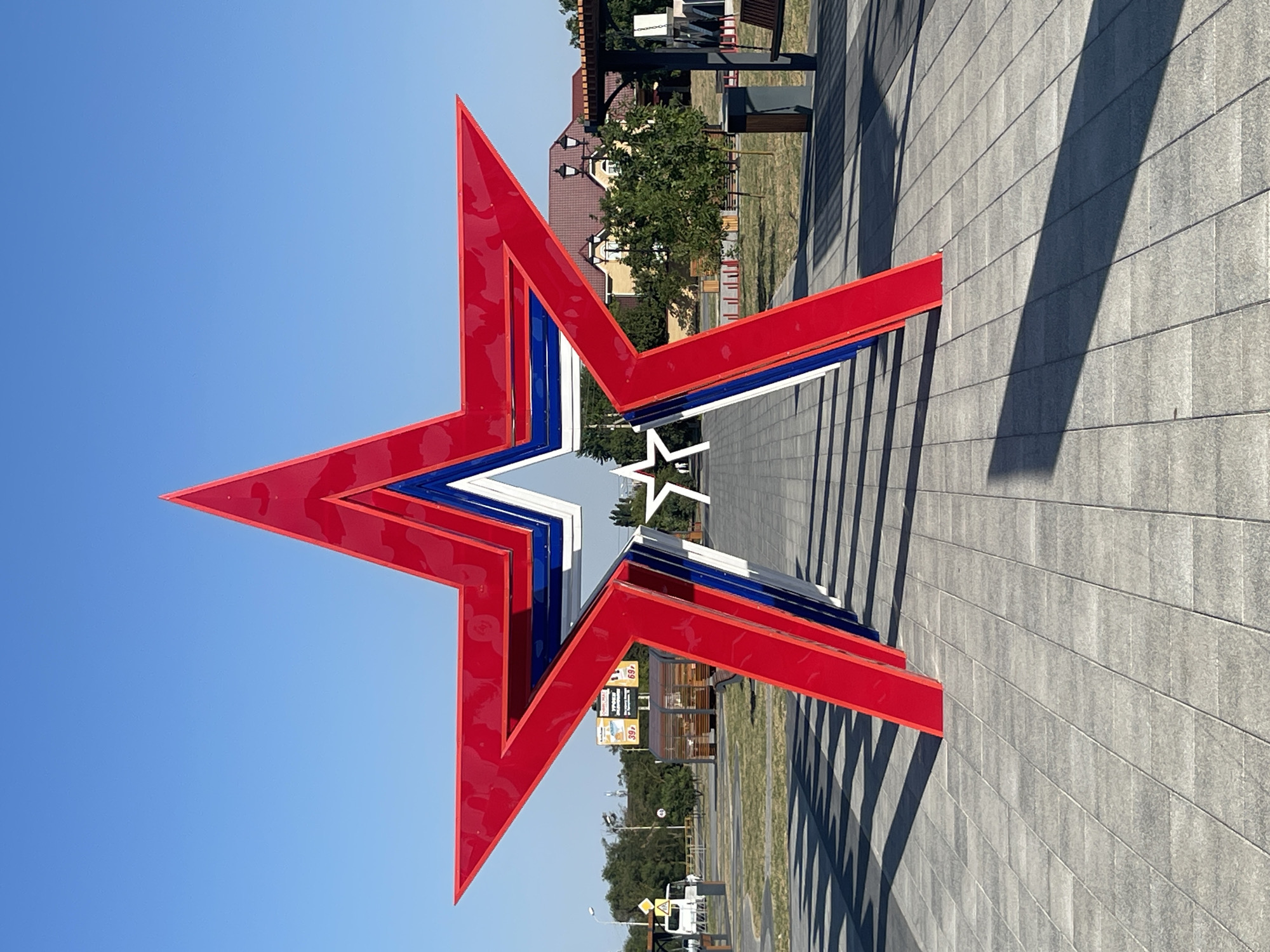 The photograph shows a large, red, white, and blue layered star sculpture in the city of Budyonnovsk, Russia. The sculpture is positioned on a paved walkway with a clear blue sky overhead. In the background, trees and buildings are visible, along with a white van. The sculpture appears to be a prominent landmark, possibly in a park or public square.