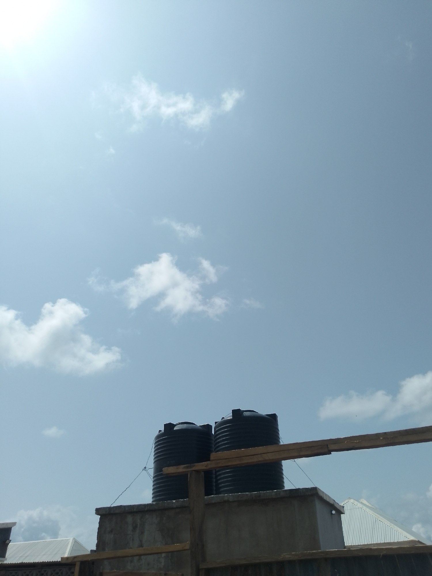The image depicts an outdoor scene featuring two large black water tanks positioned on top of a concrete structure. The tanks are cylindrical with ribbed exteriors and black caps. They are secured by thin wires. A wooden beam runs horizontally across the foreground, partially obscuring the view of the tanks. In the background, a light blue sky is visible with scattered white clouds. To the left of the concrete structure, a portion of a white corrugated roof is visible. The scene appears to be on a sunny day. The location is the city of Serti, Nigeria.