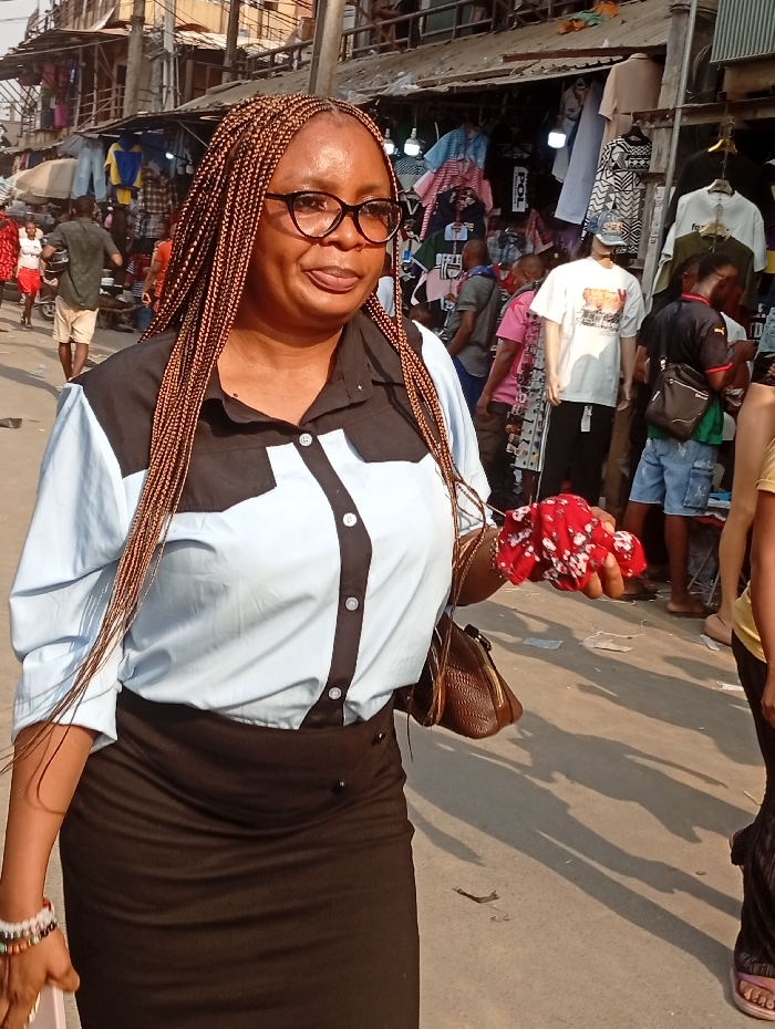 A woman with braids and glasses, wearing a black and light blue shirt and a black skirt, is walking on a sunny street with shops on either side. She carries a brown purse and a crumpled red floral cloth in her hands. The area is crowded with people and displays of clothing.