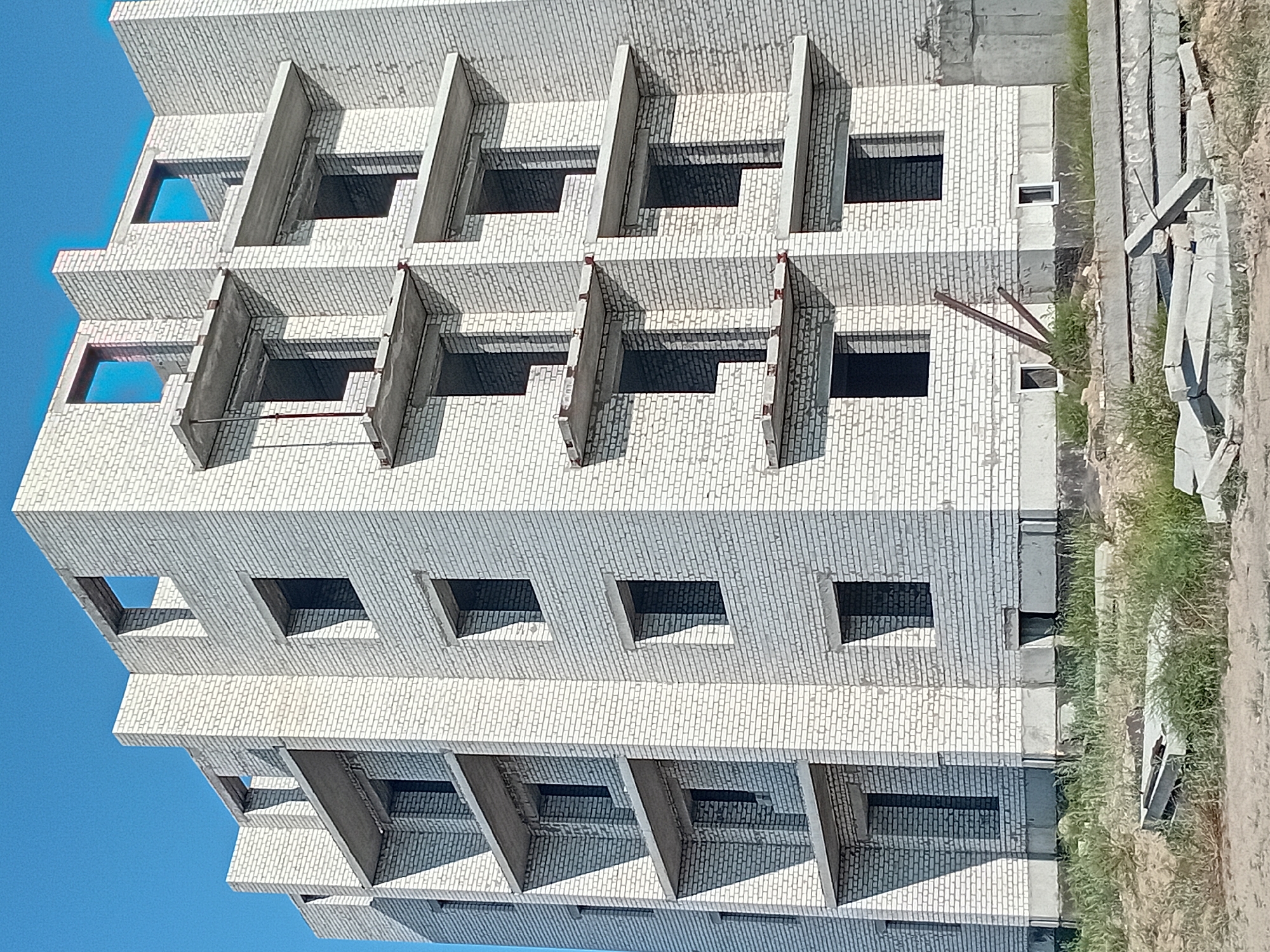 A tall, white brick apartment building is under construction against a clear blue sky. The building has multiple stories with open, unfinished window frames and concrete balconies protruding from various levels. The facade is made of evenly laid white bricks. The ground in front of the building is a mix of dry dirt and patches of green grass. Several flat concrete slabs are scattered on the ground, appearing to be construction materials. A metal pole or support beam leans against the side of the building. The scene suggests an active construction site in an urban or suburban area. The location is the city of Semey, Kazakhstan.