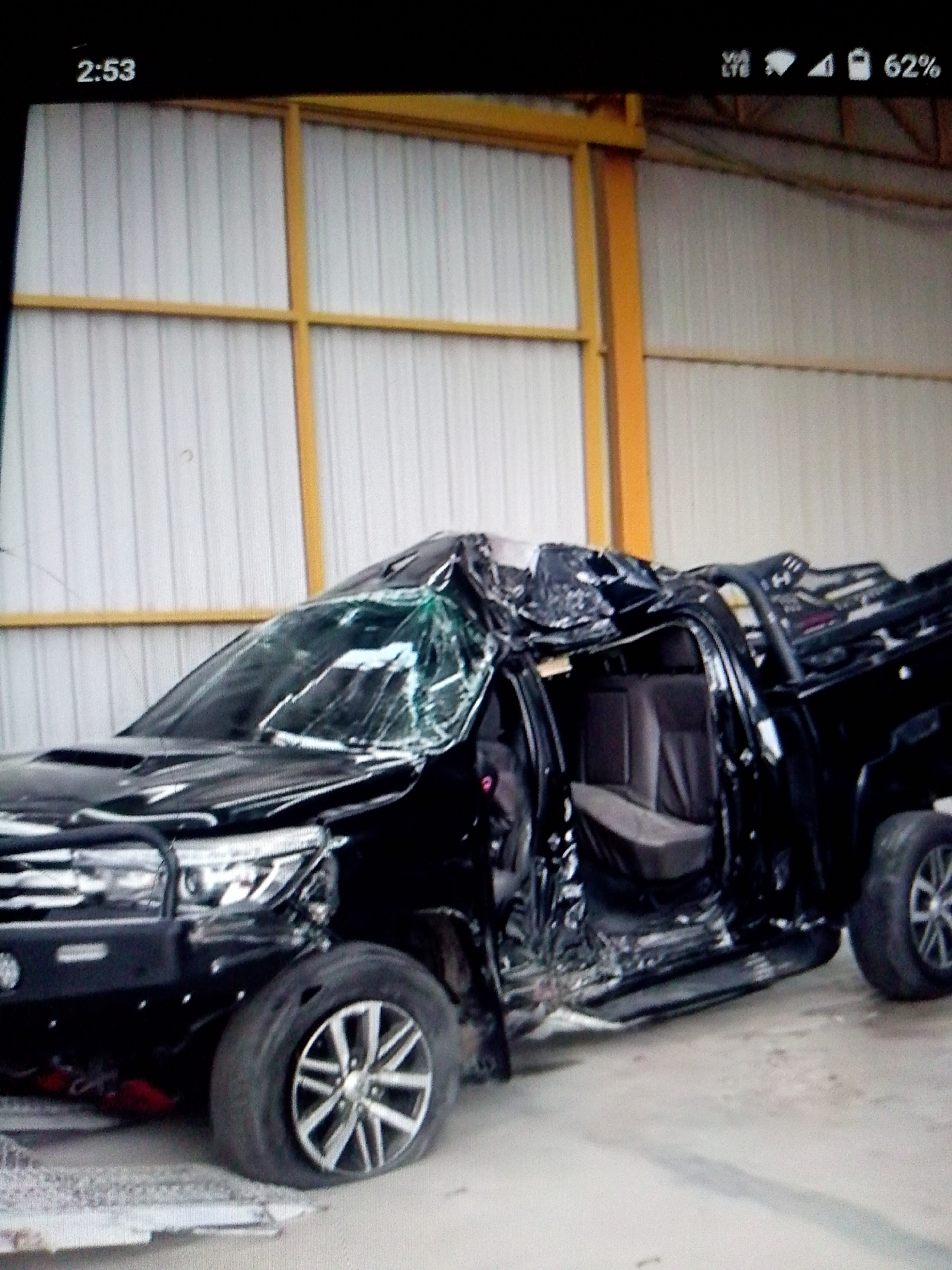 The image shows a severely damaged black pickup truck, likely a Toyota Hilux, inside a warehouse or large garage with white corrugated metal walls and yellow metal framing. The truck's windshield is shattered, the roof is crushed, and the driver's side door is missing. Both driver's side tires are flat, and the body of the truck is extensively mangled. The interior is visible and appears damaged. The overall impression is of a vehicle involved in a catastrophic accident. The time on the phone image is 2:53 and the battery is at 62%. The location is Unknown, Unknown.
