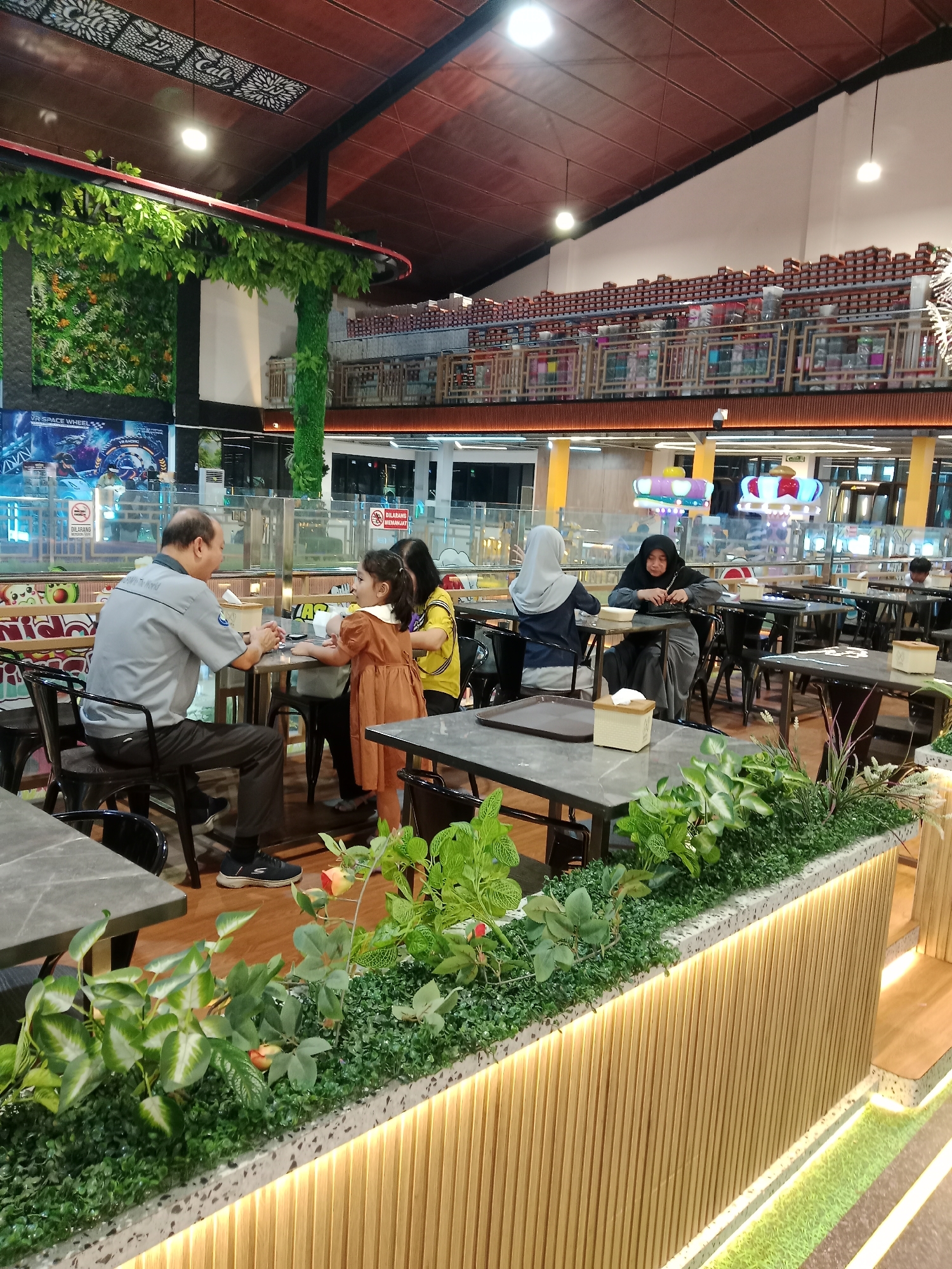 This image depicts the interior of a restaurant in Kecamatan Pati, Indonesia. The restaurant features a modern design with a high ceiling and a mezzanine level. Several individuals are seated at tables, engaged in dining and conversation. In the foreground, a man in a grey shirt is seated at a table, facing a young girl in a brown dress who appears to be interacting with him. Behind them, other patrons are visible, including women wearing headscarves. The restaurant is decorated with artificial greenery, and the tables are made of dark grey material. The overall atmosphere suggests a casual dining setting. The mezzanine level is lined with shelves displaying numerous items, and decorative lighting is visible throughout the space.