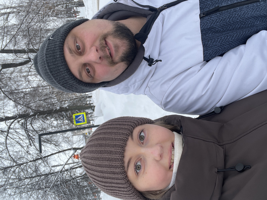 An outdoor, eye-level photograph captured in a snowy urban or park setting within Chaikovsky, Russia, features two individuals posing for the camera. The foreground is dominated by a female on the left and a male on the right, both facing forward and slightly smiling. The female wears a brown knitted beanie, a dark brown winter coat, and a light-colored scarf or neck warmer. Her eyes are light-colored. The male wears a dark gray knitted beanie and a white and dark gray winter jacket. He has a dark beard and mustache, and dark eyes. The background reveals a significant accumulation of snow covering the ground and bare deciduous trees. A yellow diamond-shaped pedestrian crossing sign with a blue square and a white walking figure is visible in the mid-ground, positioned behind the individuals. A black street lamp post stands to the left of the sign. To the far left, a blurred orange spherical object on a pole is partially visible. The scene indicates cold, winter conditions.