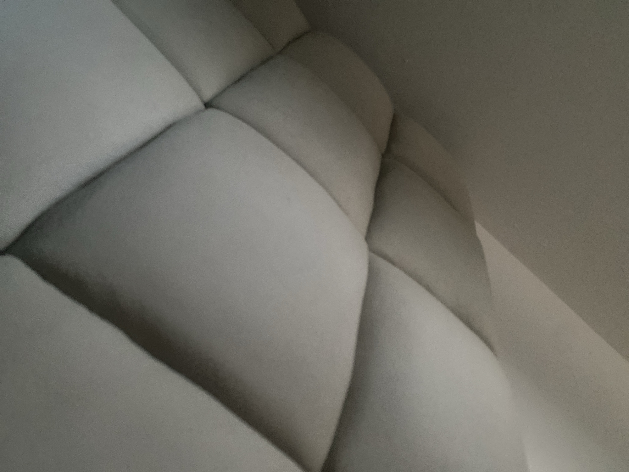 The image displays an indoor scene in the city of Jalingo, Nigeria, featuring a light-colored, upholstered surface, likely a headboard or a padded wall panel. The surface is constructed from multiple distinct, soft, rectangular segments of fabric, forming a repetitive, three-dimensional quilted or tufted pattern. Each segment is slightly raised, creating visible shadows along their edges which enhance the tactile quality and depth of the material. The fabric appears to be a muted off-white or light grey, with a subtle texture. To the left of the padded surface, a plain, unadorned wall or ceiling is visible, providing a contrasting background. No individuals, discernible objects beyond the padded structure, or specific actions are present in the frame.