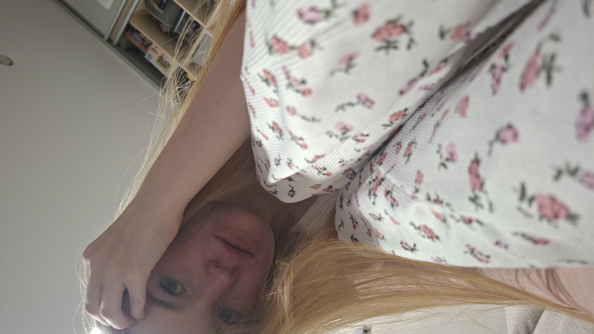 A selfie photograph from a low angle, showing a fair-skinned woman with long blonde hair and blue eyes. Her hand is touching her forehead. She is wearing a white top with pink floral print. Background shows a white ceiling and part of a cabinet with items inside. Location is unknown.