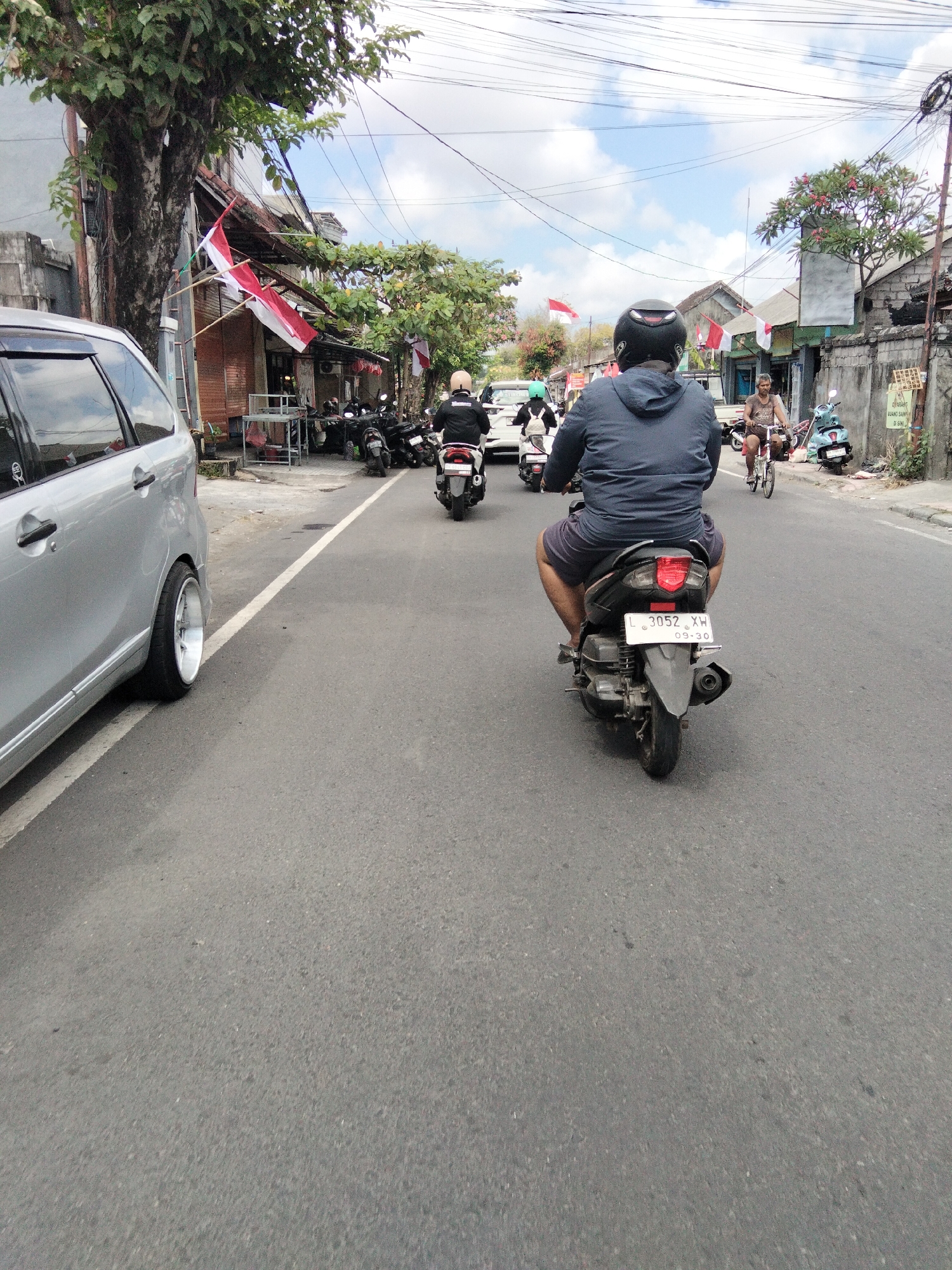 In Denpasar, Indonesia, on a paved road with a white lane marker, a silver car is parked on the left. To the right, a motorcyclist wearing a blue jacket and shorts rides a black scooter with license plate "L 3052 XH" and "09-30". Behind him, another motorcyclist in a black jacket and helmet is also on a scooter. Further up the road, two other people on scooters are visible, one wearing a green helmet. On the right side of the road, there are buildings with shops, some adorned with red and white flags, and parked motorcycles. A man on a bicycle is also seen on the right. The sky is partly cloudy.