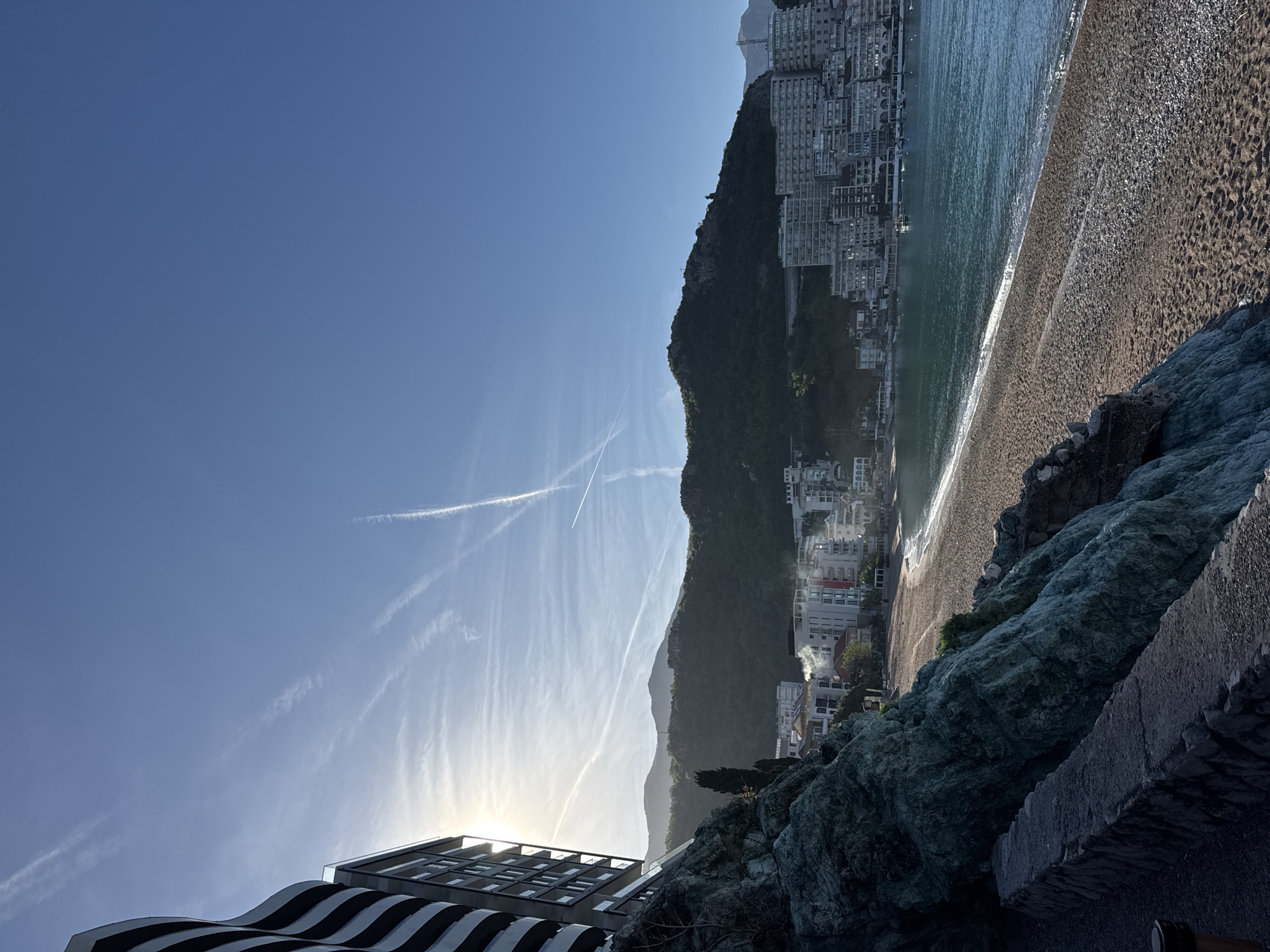 The image shows a view in Rafailovići, Montenegro, featuring the coastline, a mountain partially covered in buildings, a beach, and the sky. The beach consists of sand and pebbles, with waves breaking on the shore. Numerous high-rise buildings are built into the side of the mountain along the coast. In the foreground is a rocky cliff with some vegetation, and the corner of a building with a striped pattern. The sky is blue with contrails and the sun is visible near the horizon.