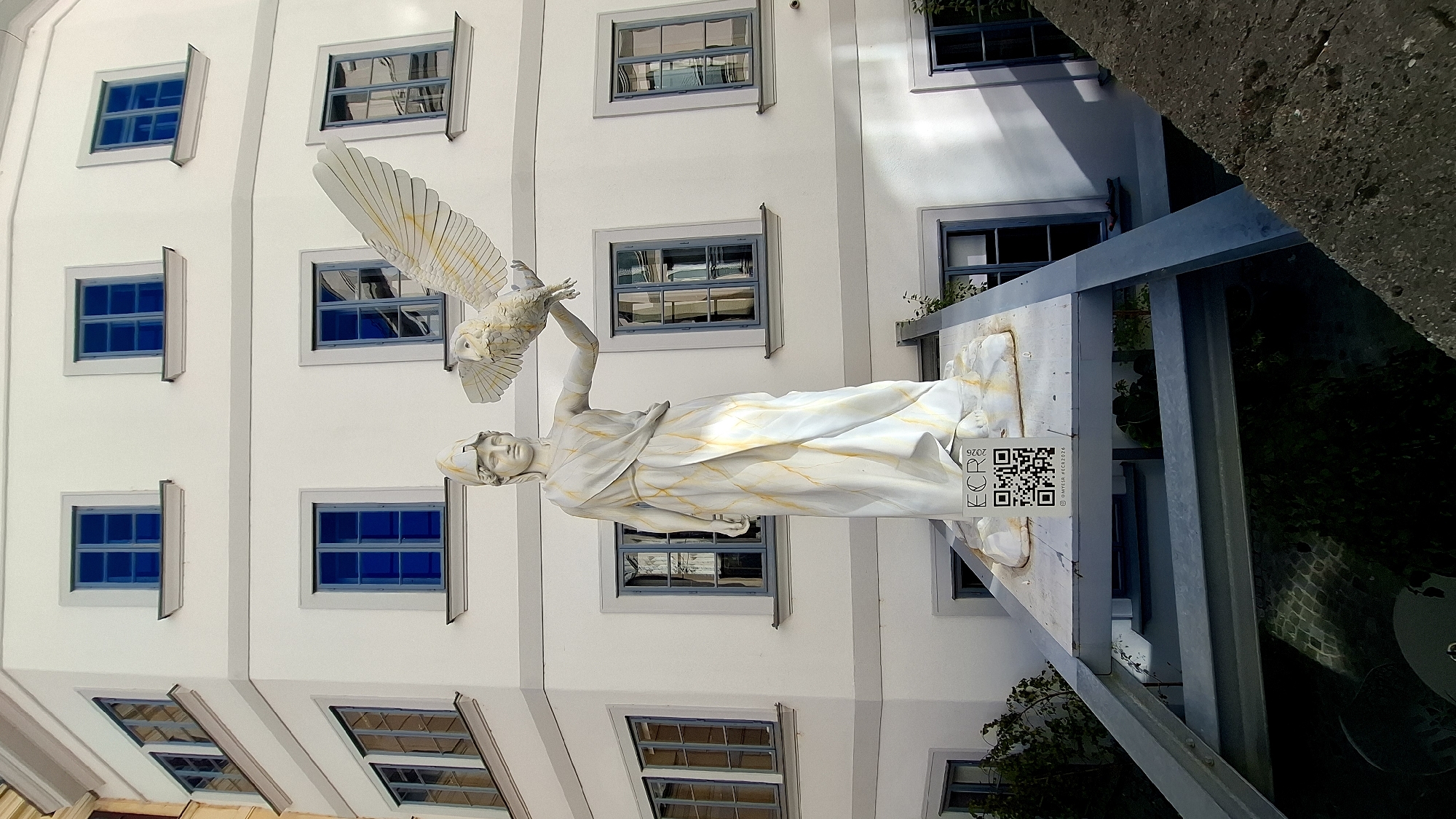 This is a medium-angle, eye-level shot of a large, white statue of a woman holding a bird with outstretched wings, set against the facade of a multi-story building in Vienna, Austria. The statue depicts a classically-inspired female figure with a helmet, draped in a flowing robe adorned with yellow accents. The bird, possibly an owl, is positioned on her outstretched hand. In front of the statue's base, a QR code and the text "ECR 2026 @MYESR #ECR2026" are visible on a white placard. The building behind the statue is white with grey trim around its windows, which are uniformly blue-paned. The scene appears to be an outdoor public space, with a metal structure supporting the statue's platform and some greenery visible below. The lighting suggests it is daytime with sunlight casting shadows.