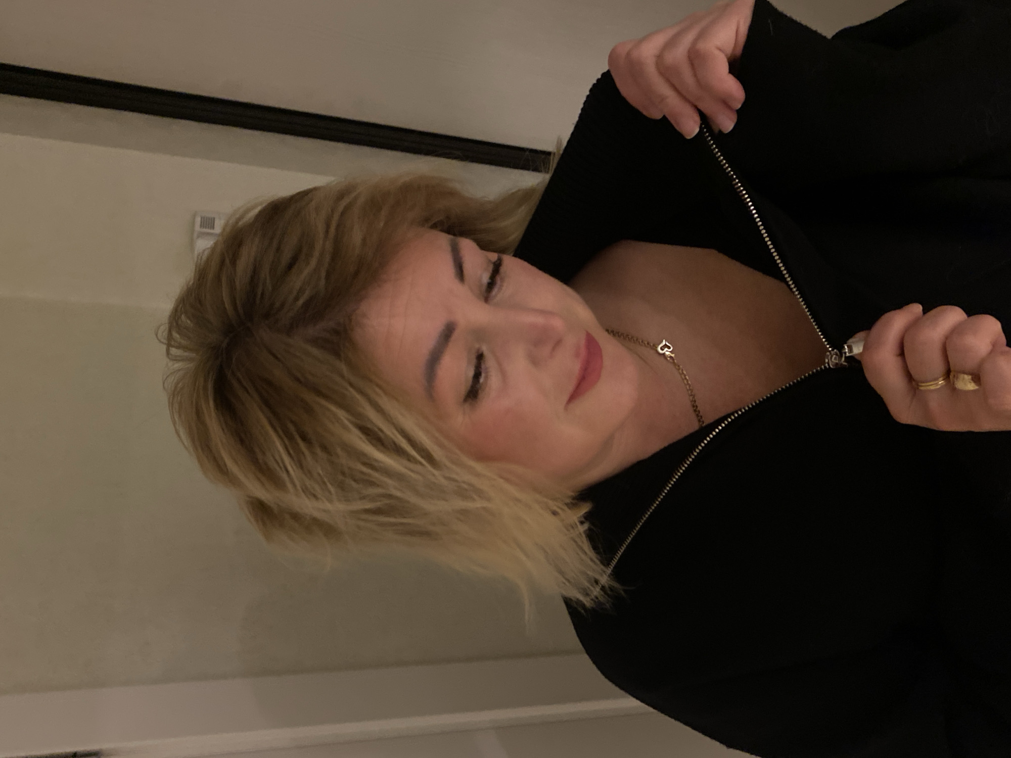 A woman with blond, shoulder-length hair is partially unzipping a black jacket. She has fair skin and is wearing a gold necklace with a heart pendant. Her right hand, with two gold rings on her fingers, is holding the zipper. The environment appears to be indoors, with a light-colored wall in the background and a dark trim along the ceiling. There is no evidence to suggest that this image was taken in Kazan, Russia.