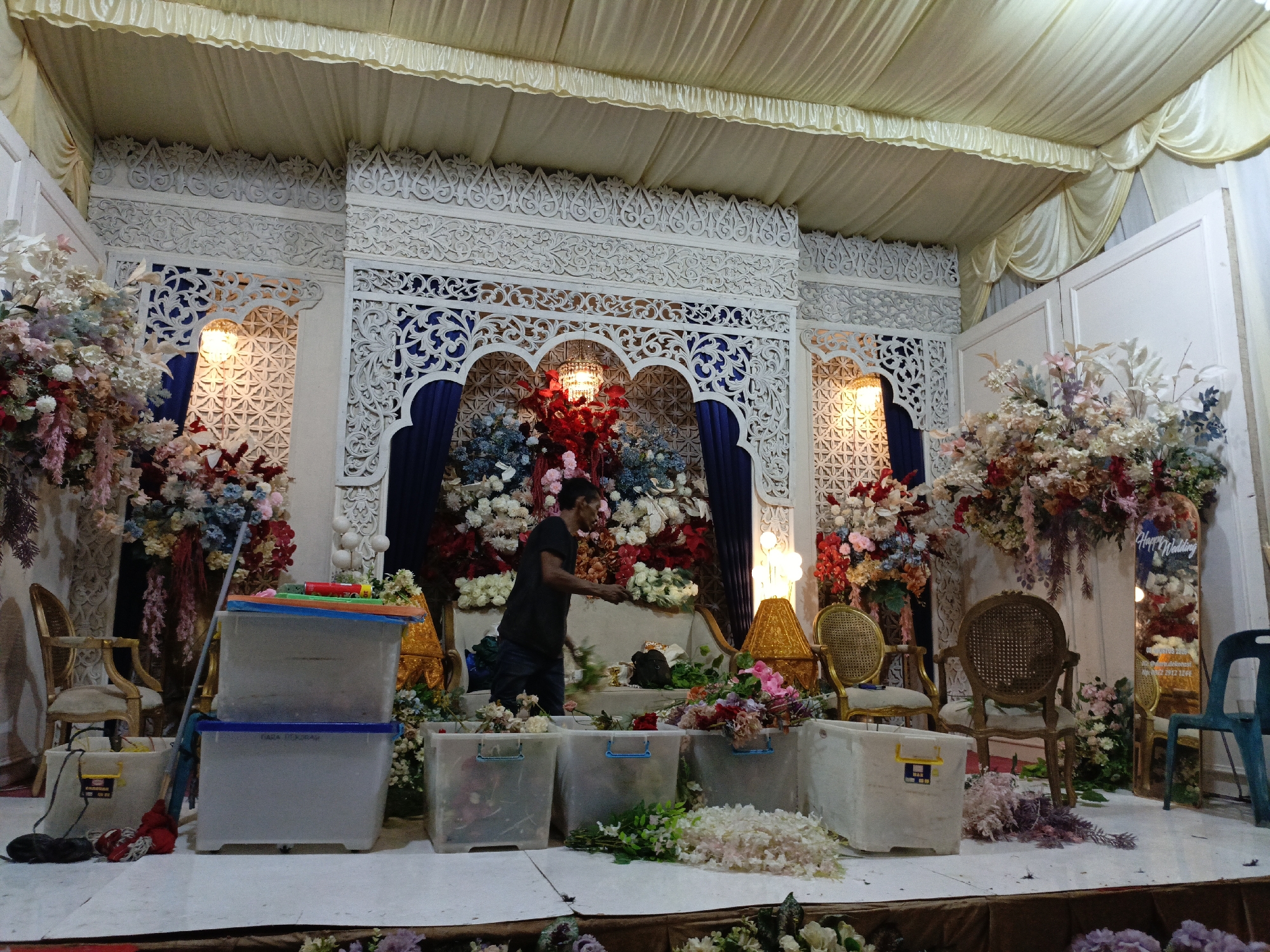 The image depicts a wedding setup indoors, likely in Pulo, Indonesia. The scene is decorated with an abundance of artificial flowers in various colors, including pink, red, blue, and white, arranged in elaborate displays. A white tented ceiling with cream-colored drapes covers the space. Intricately carved white wooden screens form the backdrop, with arched openings revealing blue drapes and floral arrangements. Golden lamps and ornate chairs are strategically placed throughout the setup.
In the foreground, several clear plastic storage bins are stacked and arranged on the floor. A man, wearing a black t-shirt and dark pants, is actively engaged in arranging flowers. He is standing in front of a sofa, holding a bouquet of flowers and appears to be working on the floral decorations. Other objects visible include a mop, a broom, and what appear to be cleaning supplies near the bins. The overall environment suggests preparations for a wedding ceremony or reception.