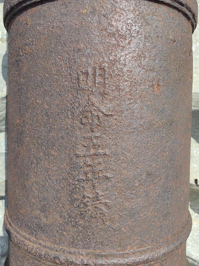 A close-up, vertical shot captures a heavily rusted cast iron object, presumed to be a section of an antique cannon barrel, located outdoors in Phú Xuân Ward, Vietnam. The cylindrical object exhibits a deeply pitted, rough, and oxidized surface, predominantly reddish-brown with darker grey patches, indicating extensive weathering. Raised characters, consistent with traditional Asian script, are vertically cast into the surface, partially obscured by corrosion but discernible. A circumferential raised band is present near the bottom of the visible section. Direct sunlight illuminates the object's right side, creating a distinct shadow line along its left. The blurred background consists of a light-colored, possibly paved or stone surface. No people, actions, or specific interactions are depicted.