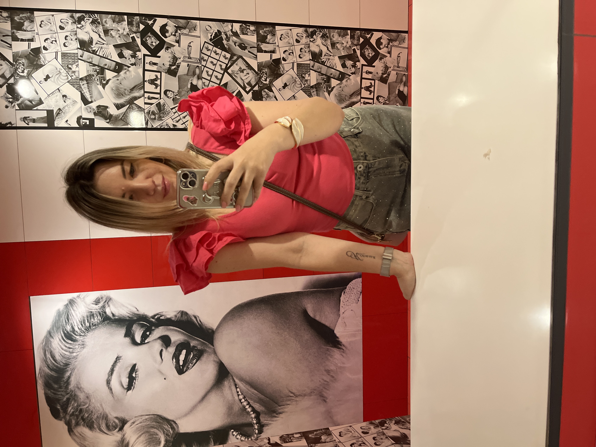 The image depicts a mirror selfie taken by an adult female in an indoor setting located in Sochi, Russia.
The woman, with blonde hair showing darker roots, is captured from approximately the waist up. She wears a bright fuchsia-colored top with pronounced ruffled short sleeves and dark gray bottoms, possibly jeans, with a subtle glitter or textured finish. A brown strap of a cross-body bag is visible across her chest. Her left forearm displays a tattoo of stylized letters, which appear to read "SOFIA" when viewed in a mirror. She also wears a light gray band on her left wrist and a white, leaf-shaped bracelet with a red cord on her right wrist. Her fingernails are painted pink. She is holding a silver-colored smartphone, consistent with an iPhone Pro model, adorned with small, reflective stickers on its case, and is looking at the phone's screen. Her right foot is elevated and rests on the white countertop in the foreground.
The background features a vibrant decor. To the left of the woman, a large, black and white poster of Marilyn Monroe is displayed. The poster, framed by a broad red border, shows Monroe from the chest up, with a pearl necklace and a visible beauty mark. To the right of the woman, the wall is covered with a collage of numerous smaller black and white photographs of various vintage celebrities and pop culture scenes, set against a white tiled surface with dark grout lines. The walls behind the counter are a mix of bright red panels and white sections. The countertop in the foreground is white and flat, with a thin black strip along its front edge, positioned above a solid red base.