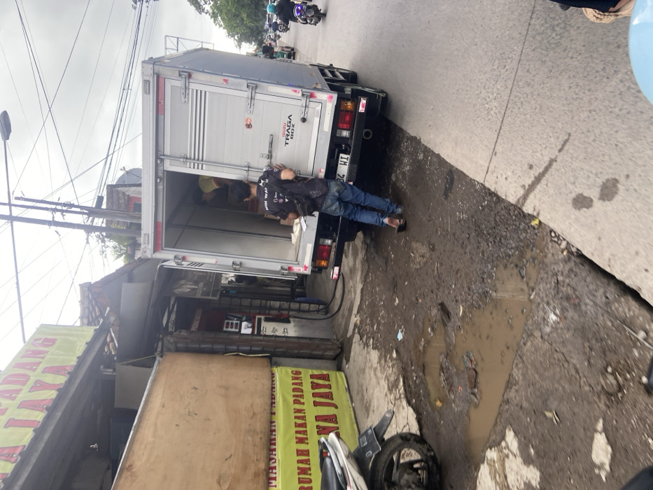 The image depicts a man in a black shirt and blue jeans standing next to a white truck. The truck is parked on the street in Baleendah, Indonesia, at the coordinates -7.021, 107.657. The truck has its rear doors open and the man is facing the camera. There is a sign in the background that reads "Rumah Makan Padang Na Jaya". There is a motorcycle in the foreground, and a puddle of water in the street in front of the truck. The street appears to be paved, but is in disrepair. The buildings in the background are a dull color. The sky is overcast and there are power lines overhead. The scene appears to be located in an urban area, possibly near a commercial district.