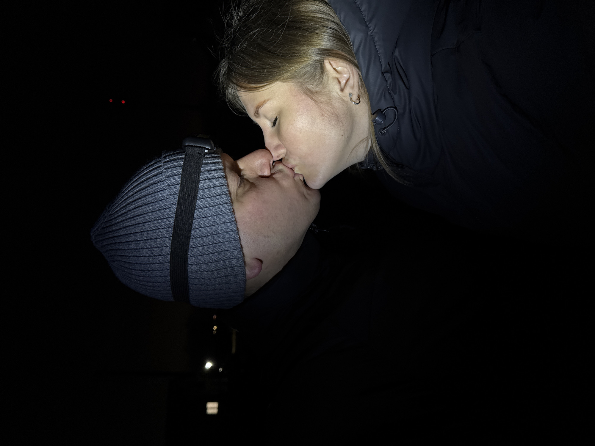 The image displays an intimate moment between two individuals kissing in a dark, likely outdoor, setting. A woman is positioned on the right, leaning in to kiss a man on the left. She has light brown hair, part of which is pulled back, and wears a dark, possibly puffy, winter jacket. A silver hoop earring is visible in her right ear, and her eyes are closed, conveying a sense of tenderness. The man wears a grey ribbed beanie with a black strap across it and a dark jacket or shirt.
The background is almost entirely black, suggesting the photograph was taken at night or in a very dimly lit environment. A few very faint, out-of-focus light sources, possibly distant streetlights or lights from buildings, are barely discernible in the deep background. One of the provided crops highlights two small, distant red dots in the otherwise black background, which are ambiguous but could be distant lights or reflections. The heavy clothing worn by both individuals suggests cool or cold weather conditions. No text is visible in the image. The scene is set in Горный, Russia.