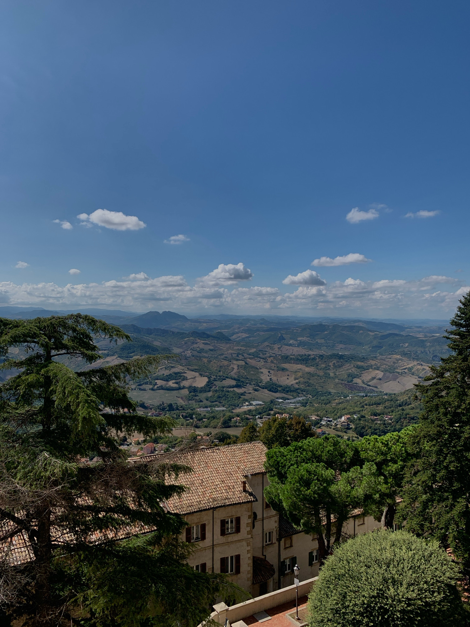The image is a high-angle outdoor shot capturing a scenic view of rolling hills and a town under a clear blue sky with scattered white clouds. In the foreground, a large evergreen tree with dense foliage is positioned to the left, its branches extending over a light-colored building with a terracotta tiled roof. The building has multiple windows with dark shutters. To the right of the building, more trees and lush green bushes are visible.
Beyond the immediate foreground, the landscape unfolds into a vista of undulating hills, dotted with fields and clusters of buildings that form a town. The distant hills are hazy, creating a layered effect. A prominent mountain range is visible in the far distance.
The scene appears to be a panoramic view from a high vantage point, likely a viewpoint in the City of San Marino, San Marino, given the architecture and the characteristic hilly terrain of the region. The lighting suggests it is a bright, sunny day. No people are visible in the image.