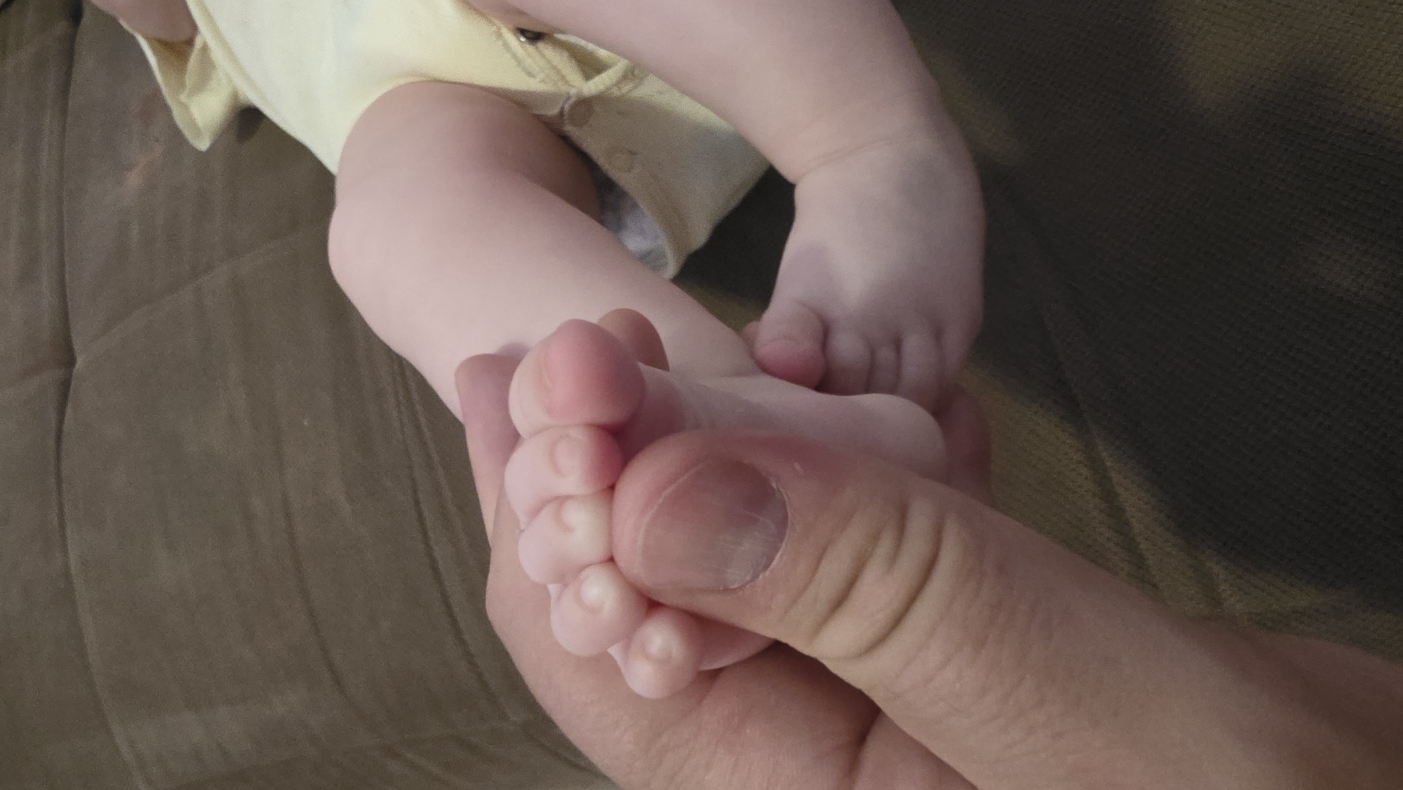 A close-up shot depicts a fair-skinned baby wearing a pale yellow onesie, lying on a brown textured surface that appears to be a sofa or couch. An adult's hand is holding the baby's tiny foot, with fingers wrapped around the toes. The skin on both baby and adult is pale. No city or location is identified in the image.
