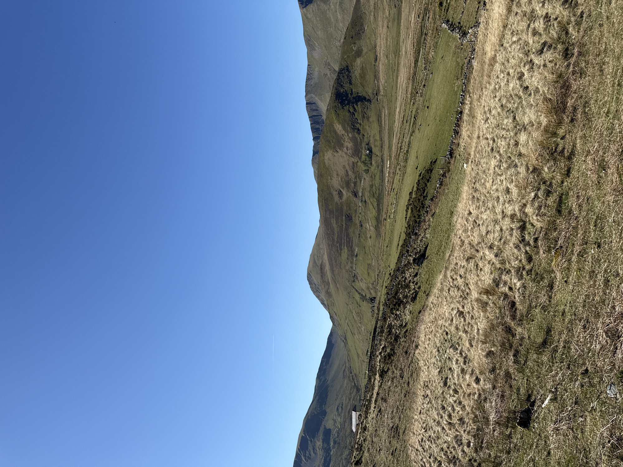 The image depicts a rural landscape in Llanberis, United Kingdom, under a clear blue sky. Dominating the scene is a mountain range with varying shades of green and brown vegetation covering its slopes. Patches of dry grass are visible in the foreground, and a stone wall runs along the mountainside. A small white structure is nestled in the mountainside.