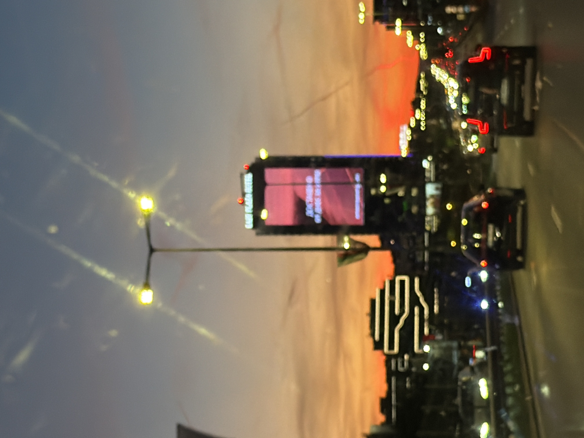 This image depicts an urban scene at dusk, likely taken from a moving vehicle. The photo is rotated, so the typical vertical orientation is horizontal. A sky gradient shifts from blue to orange. Visible elements include streetlights, an advertising billboard, buildings with illuminated signage, and traffic. The traffic consists of several cars moving along a multi-lane roadway. It's difficult to specify the exact location in Sofia, Bulgaria, due to the limited visual information.