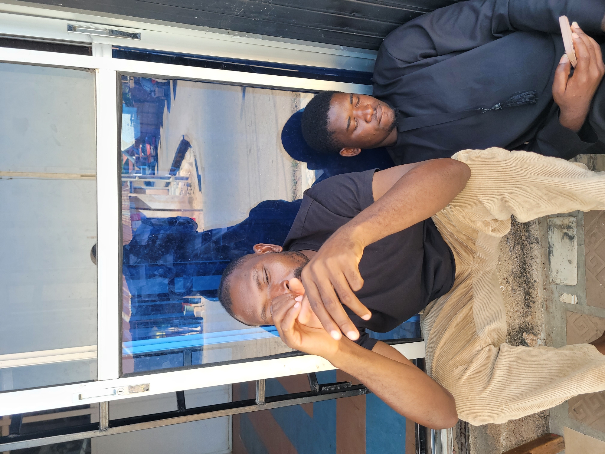 Two Black men are leaning against a glass window exterior. One man is lying with his eyes closed, wearing a black graduation gown. The other man is sitting up with his eyes closed, wearing a black t-shirt and tan pants. The reflection in the window indicates the location is a street scene in Jos, Nigeria.