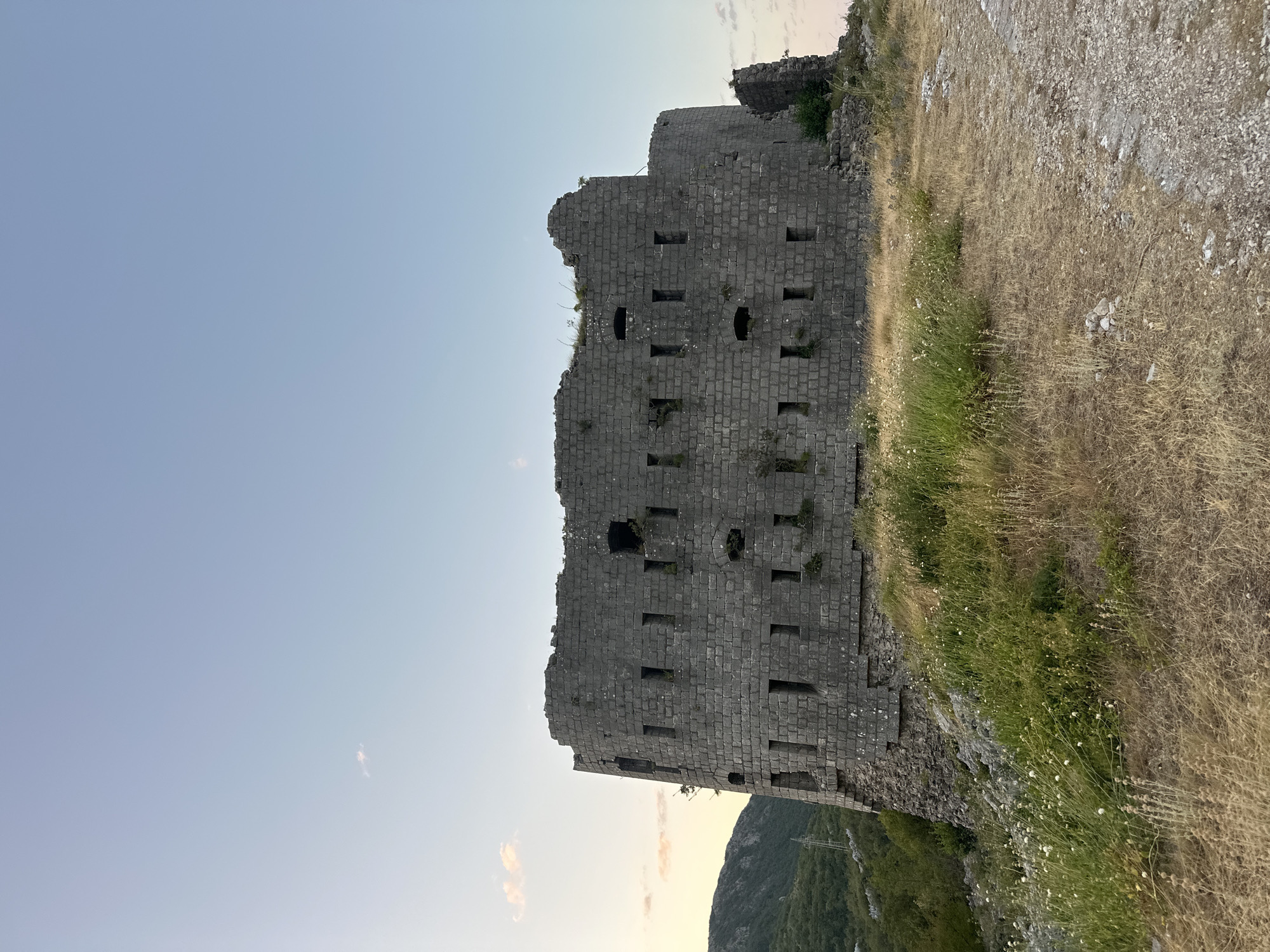 An ancient, stone fortress, likely abandoned and in a state of disrepair, stands on a grassy hillside. The fortress is constructed from large, grey stones and features numerous rectangular openings, suggestive of windows or gun emplacements. The structure has a rounded, imposing facade, with parts of the upper walls crumbled and overgrown with vegetation.
The fortress is situated in a rugged, natural environment. In the background, a lush green hillside slopes upwards, covered in trees and foliage. The sky above is a clear, pale blue, with a few faint clouds present near the horizon. The foreground consists of dry, yellow grass and rocky terrain, with a rough path made of gravel and stones leading towards the fortress.
This fortress is located in the Brajici area of Montenegro. The scene appears to be captured during the golden hour, with the soft light of dusk casting a warm glow. There are no people visible in the image, and the focus is entirely on the historic structure and its surroundings.