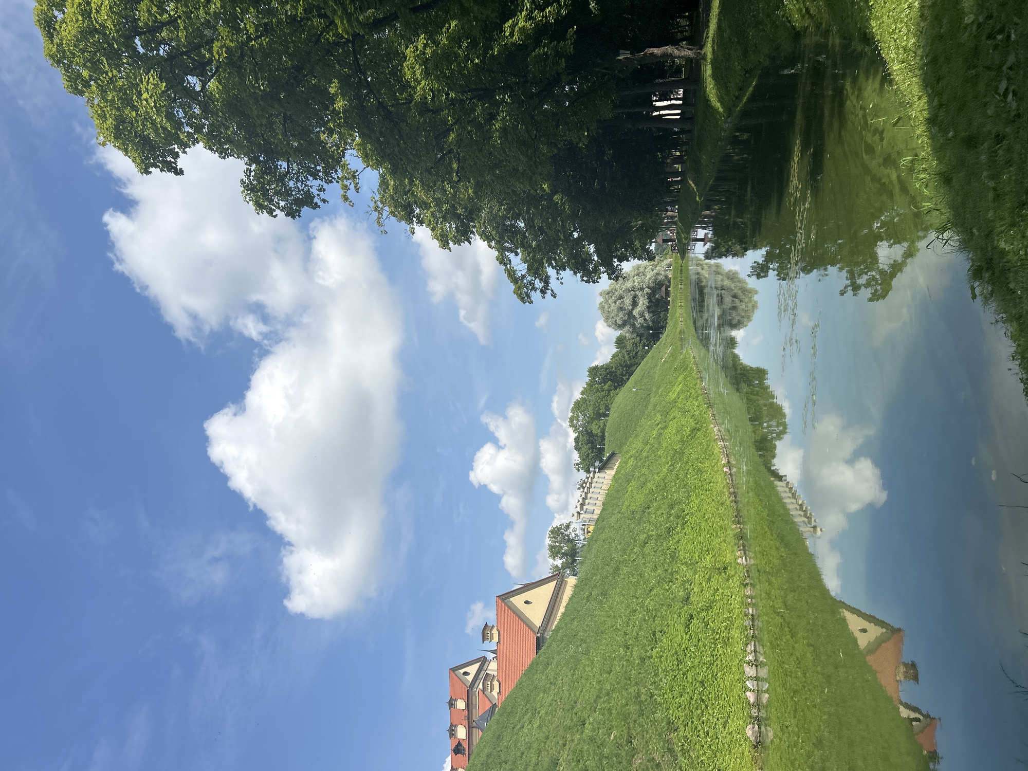 The image depicts a serene canal with its surface reflecting the sky and surrounding greenery. On the left bank, a steep, grassy hill rises, topped by a building with an orange tiled roof and arched windows. Further along the canal, on the right bank, a dense line of lush green trees forms a canopy, with a lamppost visible amongst them. The sky is a vibrant blue, dotted with fluffy white clouds. In the distance, a faint outline of a wooden structure and more trees can be seen, suggesting a park-like setting. The overall impression is one of tranquility and natural beauty within an historical context. This scene is located at Nesvizh Castle in Nesvizh, Belarus.