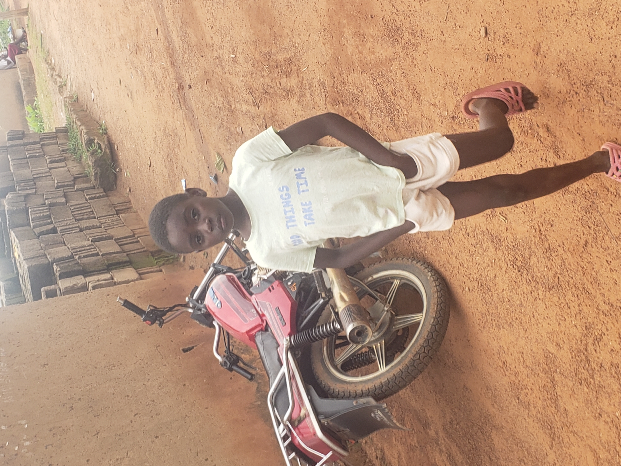 In Kachia, Nigeria, a young Black child stands beside a red motorcycle on a dirt road. The child, with dark skin and short black hair, wears a light green t-shirt with the text "GOOD THINGS TAKE TIME" in blue letters and white shorts. Their hands are in their pockets, and they are wearing pink clogs on their feet. The motorcycle, parked at an angle, has a silver exhaust pipe and wheel spokes. In the background, a wall made of stacked concrete blocks rises, with some green vegetation growing on top. The ground is a reddish-brown dirt surface with small stones. The lighting suggests it is daytime.