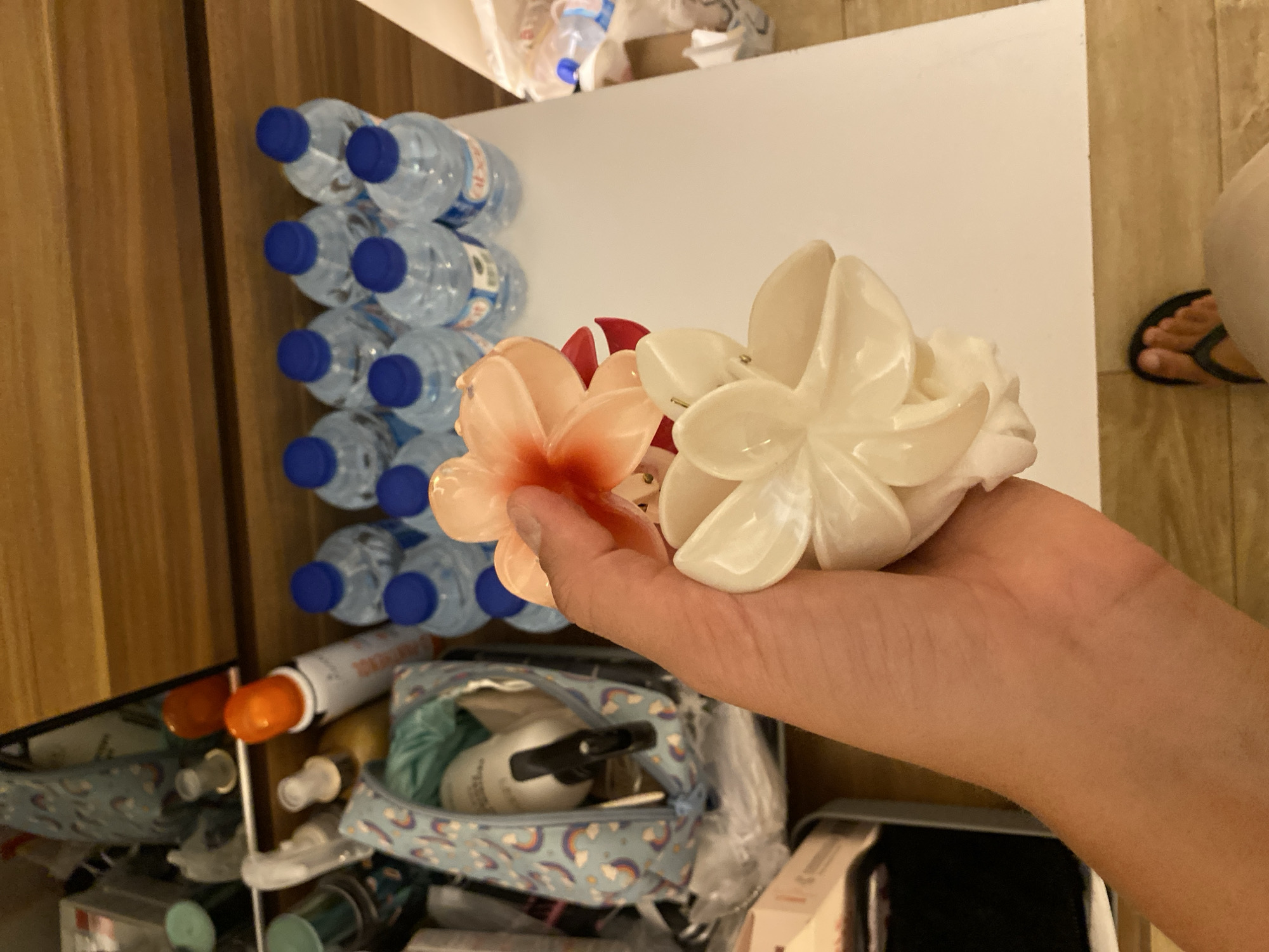 A hand is holding two floral hair clips, one peach-colored and one white. In the background, there are rows of bottled water, a cosmetic bag with a rainbow pattern, and various toiletries on a wooden surface. The scene appears to be in a hotel room.