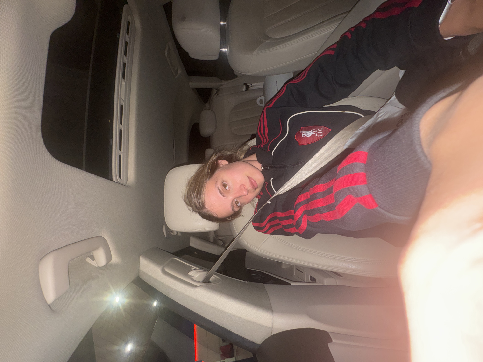 A medium shot, taken inside a car in Tbilisi, Georgia, captures a young person seated on a light-colored leather seat. The image is rotated 90 degrees to the left, showing the subject lying horizontally across the frame. The person, with light brown hair, looks directly at the camera with a neutral to slightly smiling expression. They are wearing a black tracksuit jacket with red stripes along the sleeves. On the left chest area of the jacket, a badge is visible, displaying "L.F.C." (Liverpool Football Club) and the iconic Liver bird emblem. A seatbelt diagonally crosses their torso.
The car's interior is predominantly light-colored, featuring cream or beige leather seats, a matching headliner, and door panels. Visible details include a roof-mounted air vent, a grab handle, and reflections of external lights (possibly streetlights or building lights) in the car's windows, suggesting it is evening or night time. The overall lighting inside the car is dim, with some brighter reflections and flares, possibly from a camera flash or an external light source, especially towards the bottom right. The general setting indicates a modern and well-maintained vehicle.