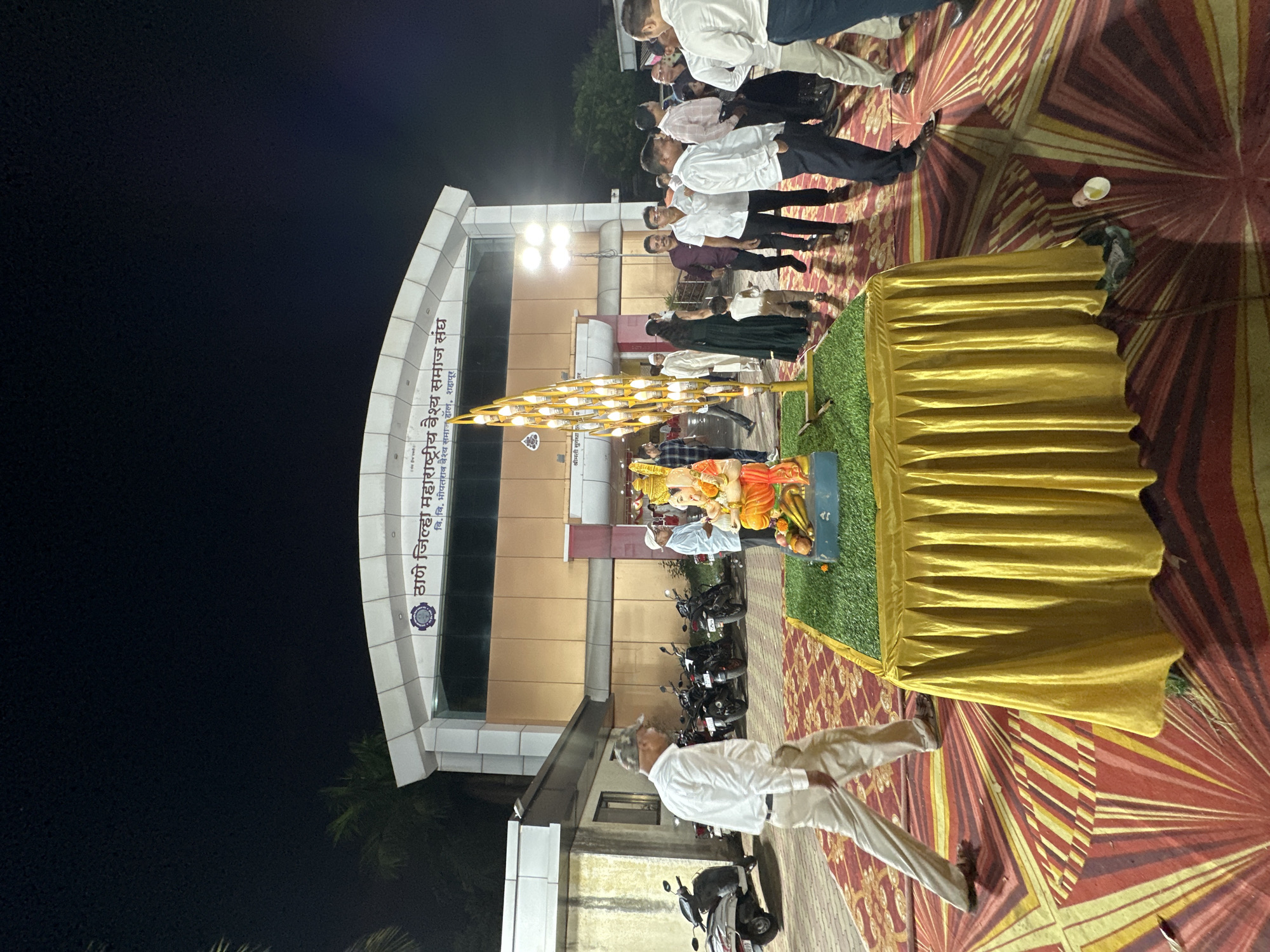 Here is a description of the image:
The image captures an outdoor event in Shahapur, India, specifically in front of a building identified as "Thane Jilha Maharashtra Vaisya Samaj Sangh" and "V.V. Bhopatrao Vaishya Samaj Hall". Several people are visible, some standing in a group. A statue of a deity is positioned on a platform draped with a golden cloth. The ground is covered with a patterned carpet. Several motorcycles are parked near the building. The sky is dark, suggesting it is nighttime. A man in light-colored clothing is walking across the lower portion of the frame.