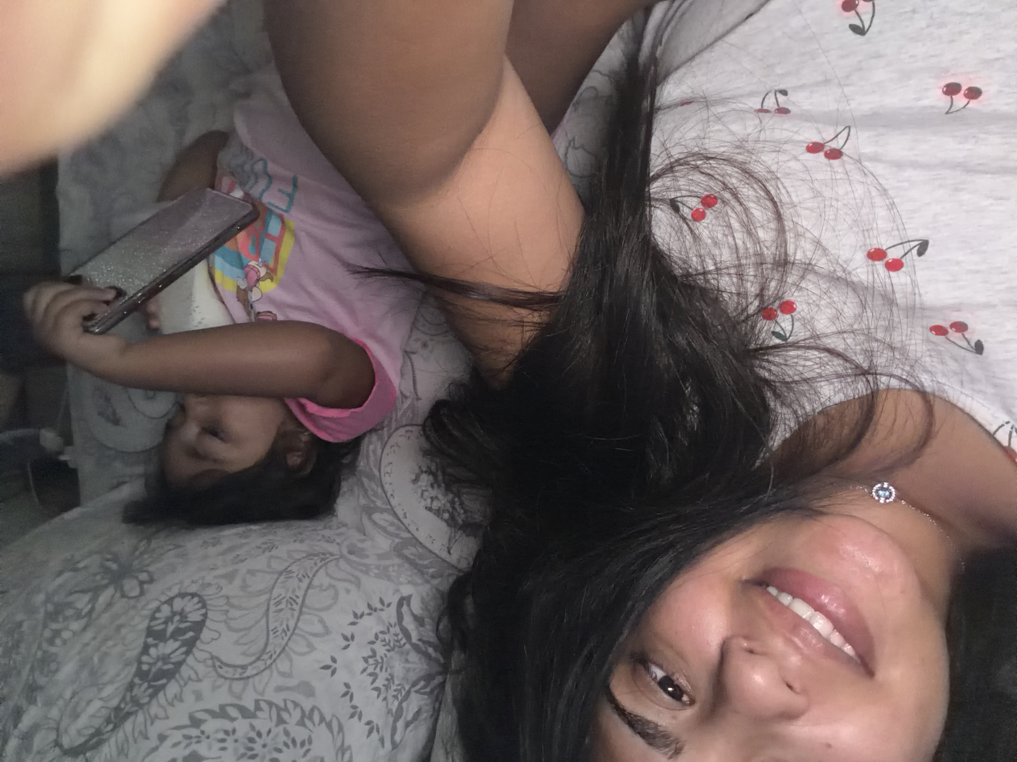 The image shows a close-up of two individuals, a woman and a child, lying down in bed. The woman, in the foreground, has dark hair and is smiling at the camera. She is wearing a white t-shirt with a cherry pattern. A small, delicate necklace with a blue and silver pendant is visible on her neck. Behind her, a young child is lying down, holding a sparkly phone. The child is wearing a pink shirt with a cartoon character on it. Both are on a bed with patterned grey and white bedding. The scene appears to be a casual, domestic setting. There is no indication of the specific location within Duisburg, Germany.