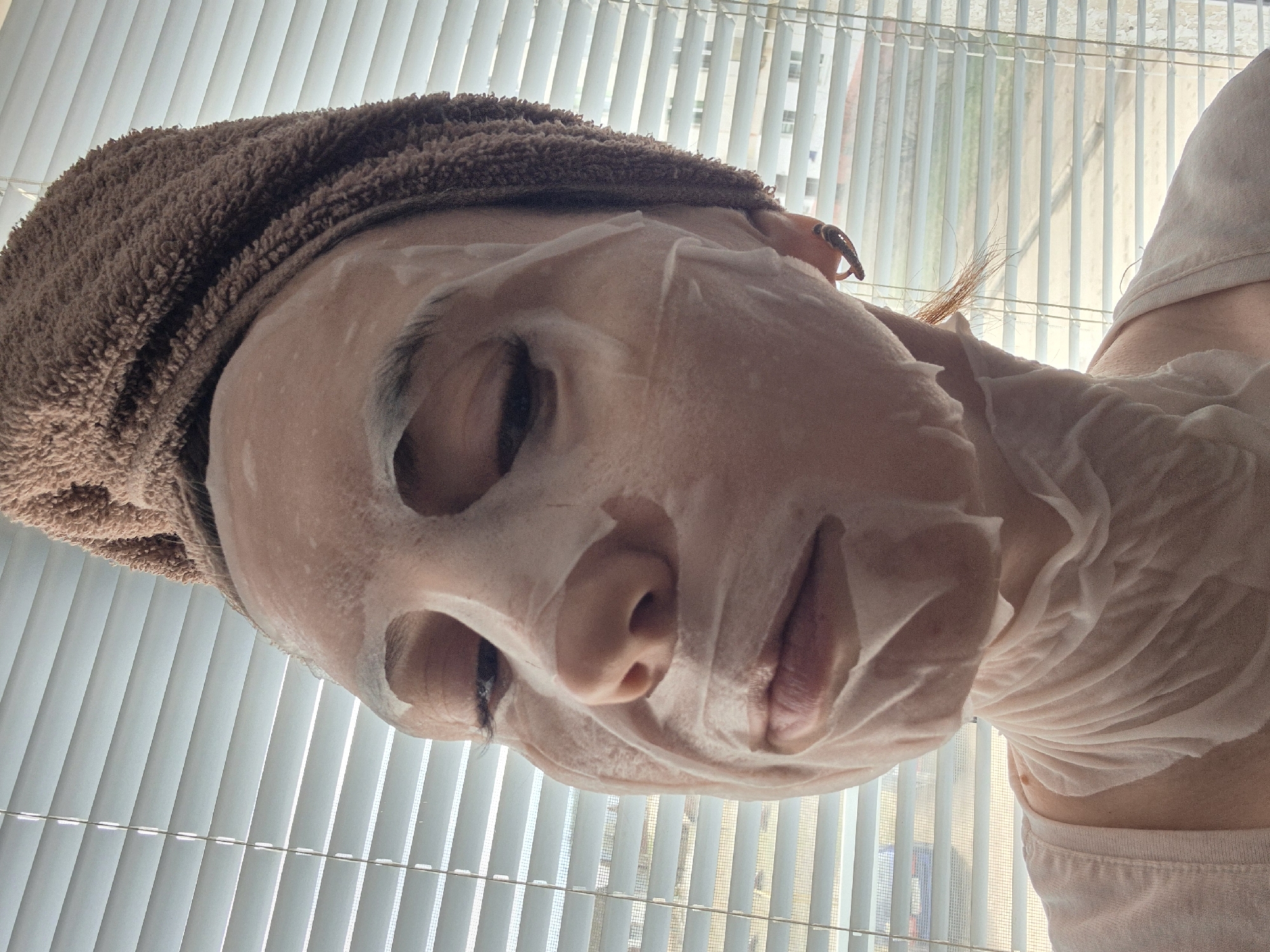 A medium close-up shot depicts a person's face obscured by a sheet mask, with a brown towel wrapped around their head. The person appears to be looking down at the camera.
Behind the person, a window with closed white blinds is visible, suggesting an indoor setting. Through the blinds, hints of buildings can be seen in the distance.
Without further information or context, it's impossible to definitively determine if this scene is located in the city of Chaikovsky, Russia.