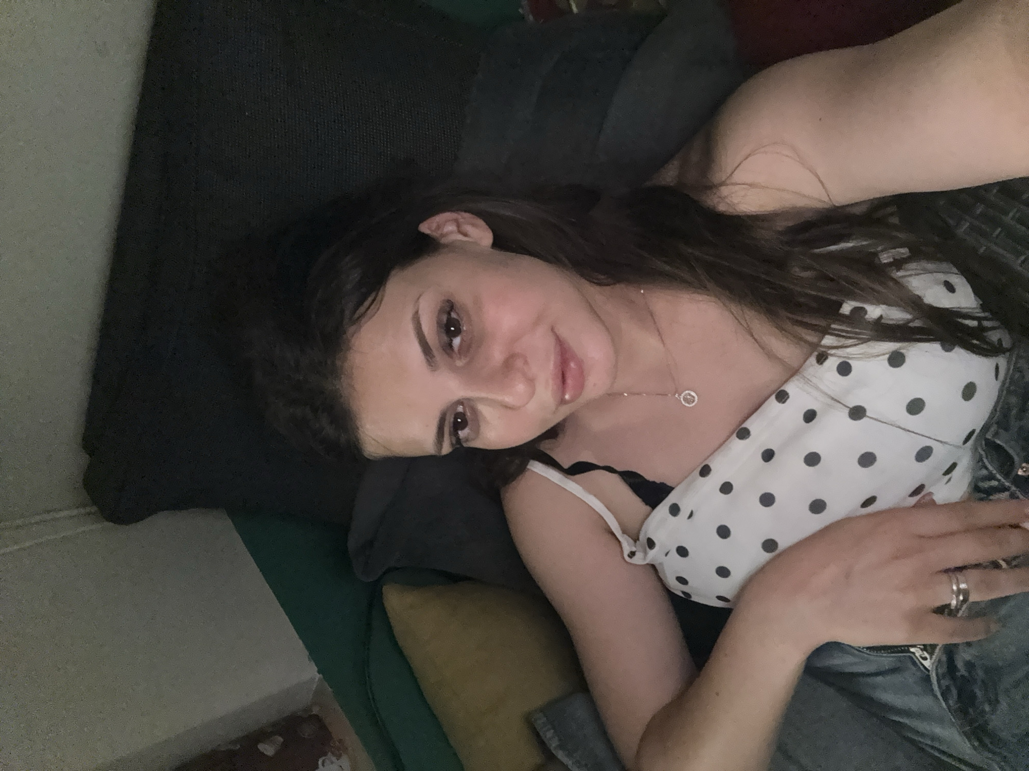The image shows a young woman lying on a couch, looking at the camera. She has long, dark hair and is wearing a white top with black polka dots and a silver necklace. Her left arm is resting on her chest, and she is wearing two rings on her left hand. The couch appears to be in a dimly lit room, with pillows visible behind her. There are no discernible Plovdiv, Bulgaria landmarks or features in the image.