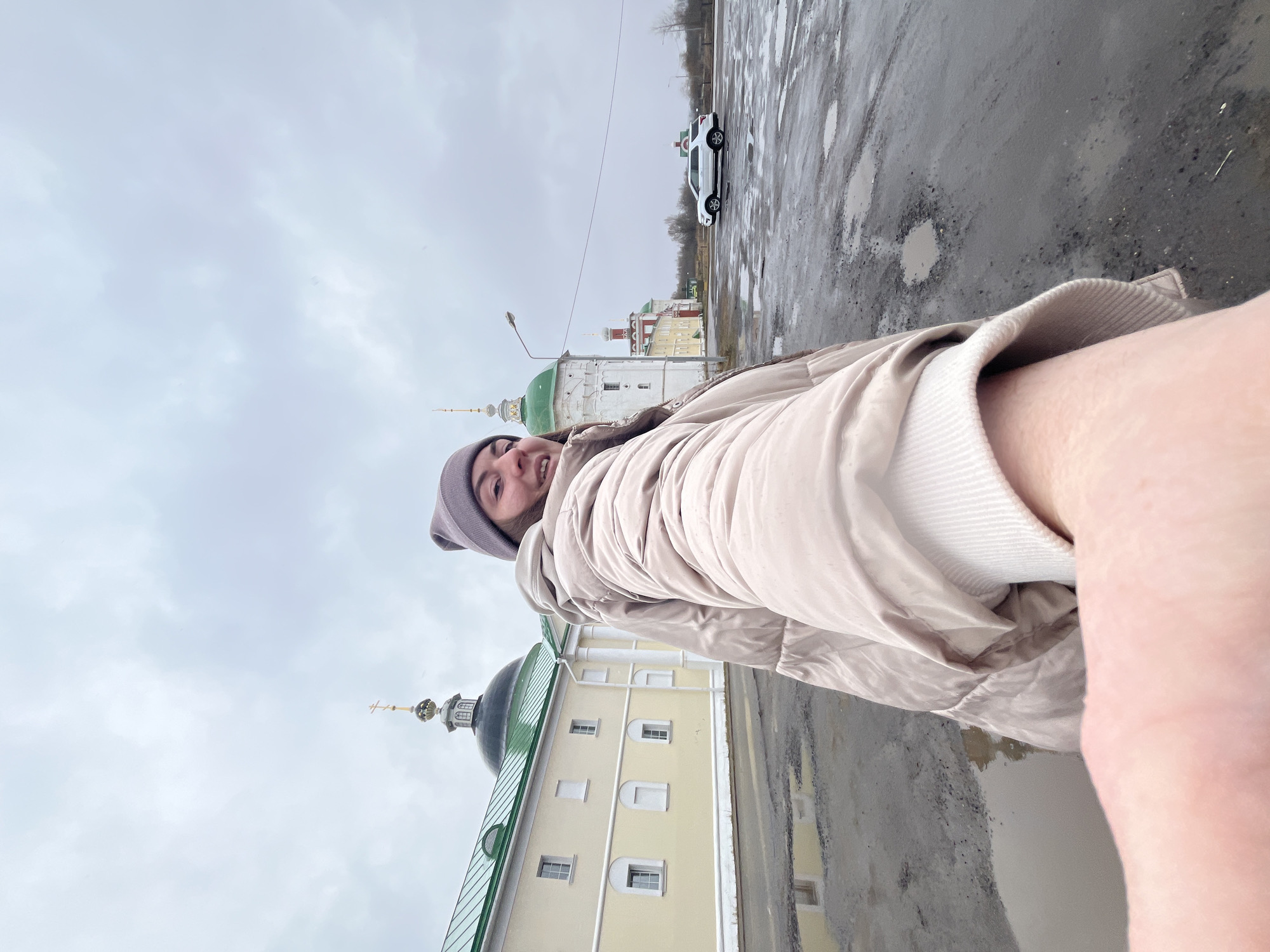 A vertical, medium shot shows a person in the city of Луговой, Russia, with a religious building in the background and a parked car in the distance. The person, who is at a tilted angle, is wearing a gray beanie and a light brown jacket. The building in the background has a pale yellow façade with symmetrical windows and a green domed roof topped with a cross. The area is wet with puddles, indicating recent rain. The sky is overcast and gray. A white car is parked on a paved area in the background, suggesting a street scene.