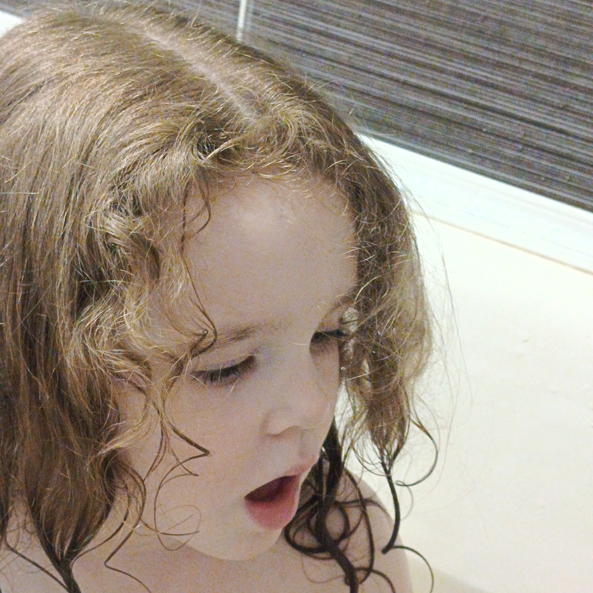 The image depicts a young girl with wet, blonde, curly hair looking downward with her mouth open. She appears to be in a bathroom, as evidenced by the tile in the background and what may be the edge of a bathtub in the lower part of the frame. The image is a close-up of her face. The location Марковский, Russia is not detectable from the image content.