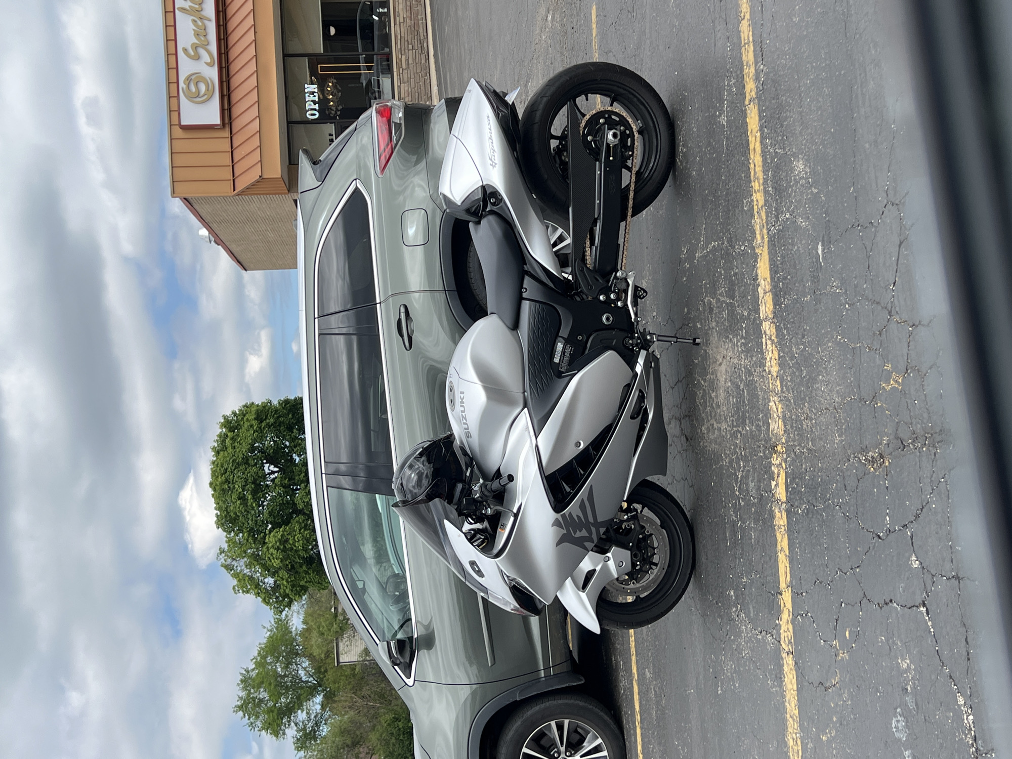 The image shows a gray Suzuki Hayabusa motorcycle parked next to a gray Nissan Rogue SUV in a parking lot. The parking lot has yellow lines painted on cracked asphalt. In the background, there's a building with a sign that reads "Sushi" and an "Open" sign visible in the window. A tree is visible behind the SUV. The location is identified as the city of Unknown, Unknown.