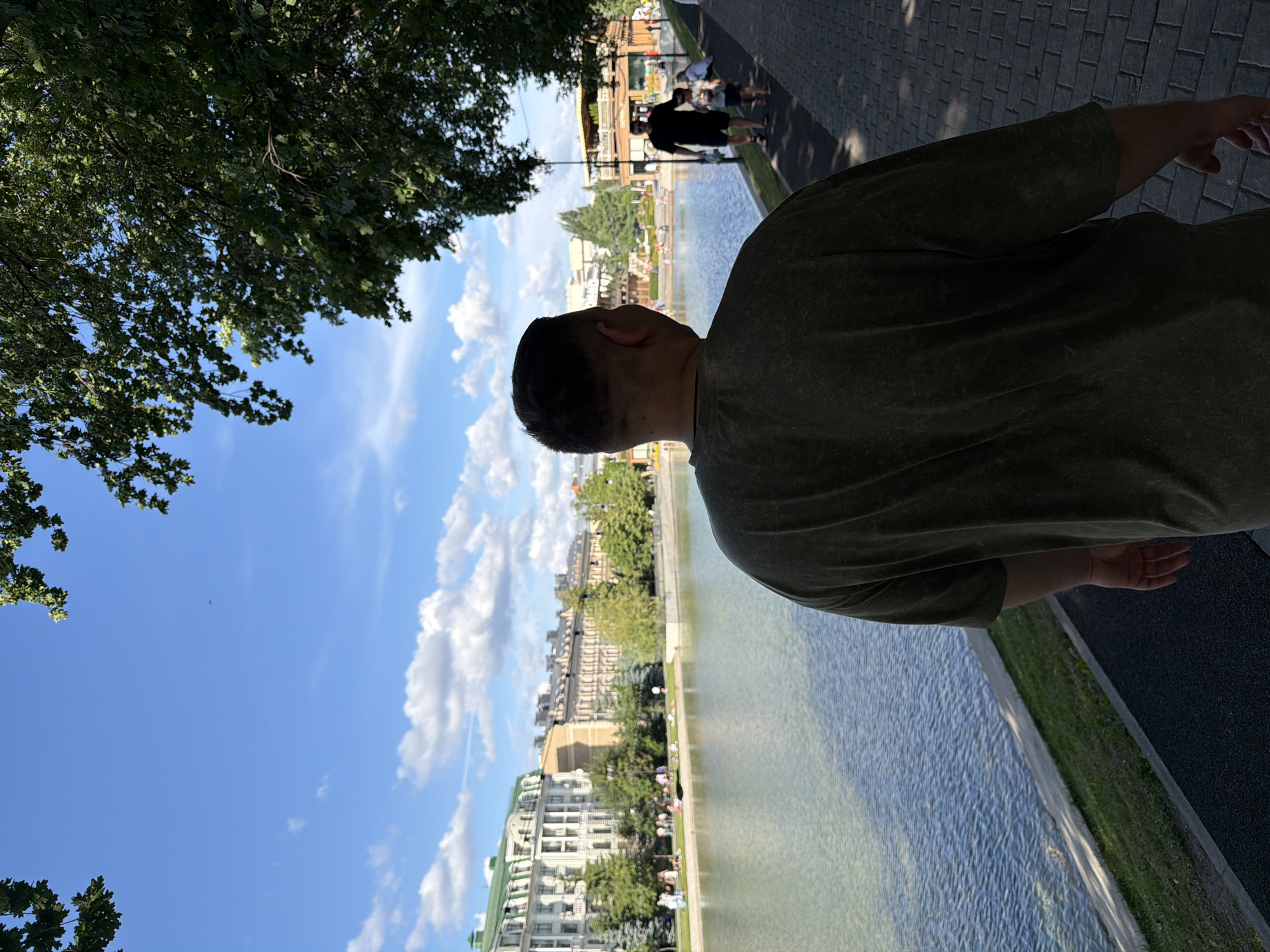 This image depicts a sunny day in Kazan, Russia, along a waterfront promenade. In the immediate foreground, a person with short, dark hair, viewed from behind, wears a loose-fitting dark green t-shirt and walks on a dark paved path. To the left of the path, a calm body of water stretches across the midground, reflecting the sky and distant structures.
On the far bank of the water, several multi-story buildings with classical architectural elements are visible, some featuring green roofs. A row of trees, including both coniferous and deciduous types, lines the far bank, interspersed with green lawn areas where several indistinct figures are scattered, appearing to relax or walk.
To the right of the central subject, a wide paved path, constructed from dark, rectangular pavers, extends into the distance. This path is partially shaded by dense foliage from large trees, whose branches with green leaves frame the upper right and upper left corners of the image. Sunlight casts dappled shadows on the path. On this right path, a man in a dark t-shirt and shorts walks towards the viewer, holding the hand of a small child. Another adult figure is partially visible further down the path.
The sky above is a clear, bright blue with scattered white cumulus clouds. High in the sky, a small, dark outline of an airplane is discernible.