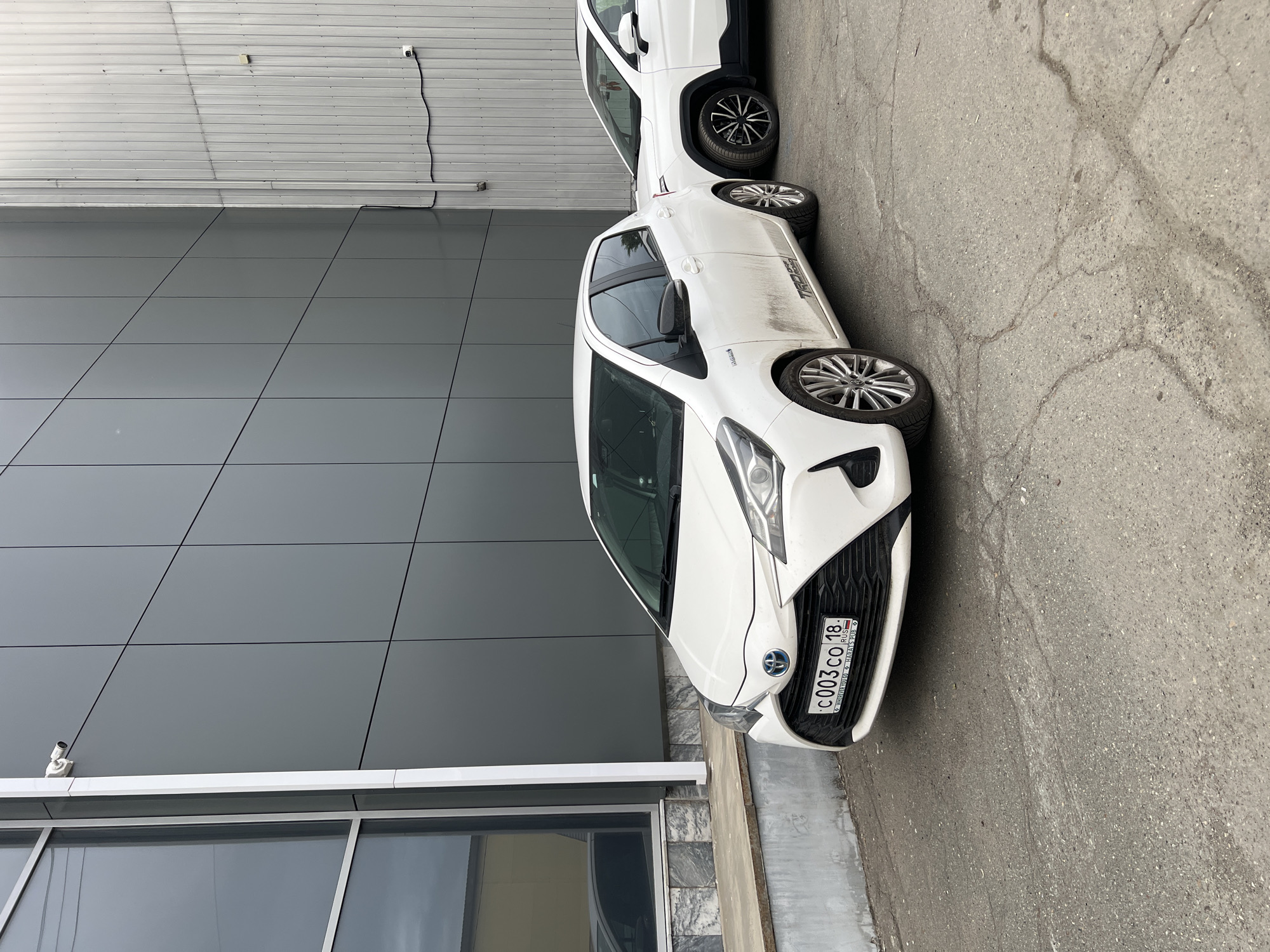 The image presents an outdoor scene in Izhevsk, Udmurt Republic, Russia, as evidenced by the "18 RUS" country code on the visible license plate. Two white vehicles are parked on cracked asphalt pavement, adjacent to the exterior walls of two commercial or industrial buildings.
In the foreground, a white Toyota Yaris Hybrid hatchback is parked, facing slightly left. The vehicle's lower body panels are visibly dirty. It is equipped with multi-spoke alloy wheels and features a "HYBRID" badge on the front fender and a "TRD pro road" decal on the lower rear passenger door. The front license plate displays the registration number "c003co 18 RUS" and includes the text "WHATEVER YOU GO HARATS PUB" in smaller print below the main number.
Partially visible on the right, a second white crossover or SUV is parked. This vehicle has black fender flares and multi-spoke wheels with black and silver elements.
The building on the left features a modern dark grey panel facade with grid-like segmentation and large, reflective window sections framed in white. A white, bullet-style security camera is mounted on the upper portion of this building's corner. The building on the right has a lighter grey, vertical corrugated metal siding. A vertical conduit runs down the seam where the two buildings meet. A second security camera, also white, is mounted lower on the corrugated wall, with a black cable extending from it. The ground in the immediate vicinity of the left building includes a concrete foundation and a raised ledge. No individuals are present in the image. The lighting suggests an overcast sky.