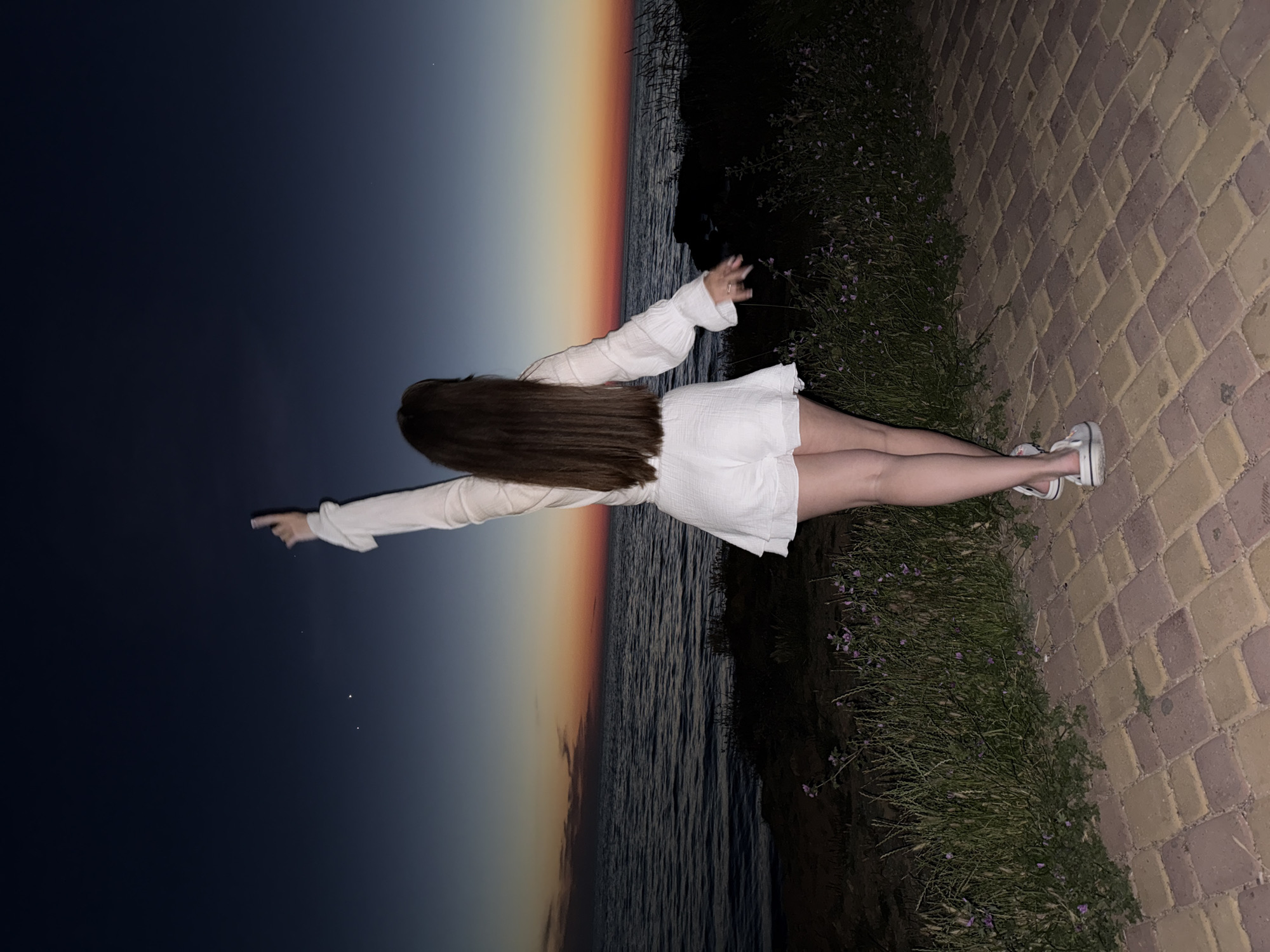 A woman stands with her back to the camera, facing the sea at dusk. She wears a white long-sleeved top and white shorts. Her right arm is raised, pointing upwards, and her left arm is extended to her side. The sky displays a gradient of colors, transitioning from deep blue at the top to orange and red near the horizon, where the sun has recently set. The sea is calm, with gentle waves. The foreground is a paved walkway made of brick-like stones in shades of brown and yellow. To the left of the walkway, there is a strip of green grass and some small purple flowers. The overall atmosphere is serene and captures a moment of appreciation for the natural beauty of the sunset. The location is unknown.