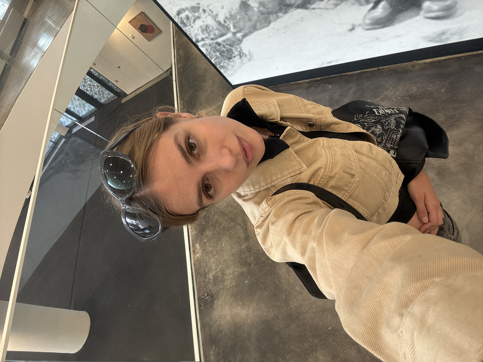 A tilted medium shot shows a woman with light brown hair and sunglasses on her head, wearing a beige jacket and a black backpack, as she stands in what appears to be a museum. The background includes a large black-and-white image on the wall and architectural details of the building. The image was taken in Yekaterinburg, Russia.
