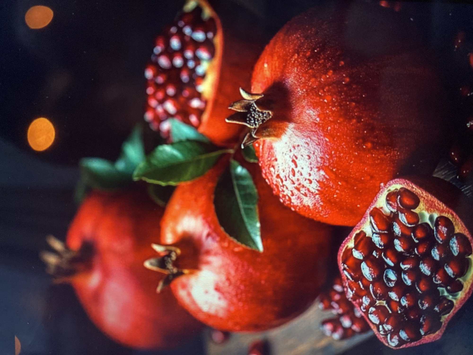 A close-up shot focuses on a cluster of pomegranates, with three whole fruits and one halved. The pomegranates are a deep, rich red, glistening with water droplets, suggesting they have been recently washed or are naturally dewy. One pomegranate is prominently displayed in the foreground, with its stem end, featuring small, dark seeds, clearly visible. To its left, another whole pomegranate is partially obscured. In the background, a third whole pomegranate is visible, and to the right, a halved pomegranate reveals its vibrant interior, packed with juicy, ruby-red arils. A few green leaves are interspersed among the fruits, adding a touch of natural color. The lighting is dramatic and moody, with soft, warm bokeh effects in the background, possibly from out-of-focus lights, creating an intimate and possibly evening ambiance. The overall impression is one of freshness and abundance. There is no visible text in the image.
