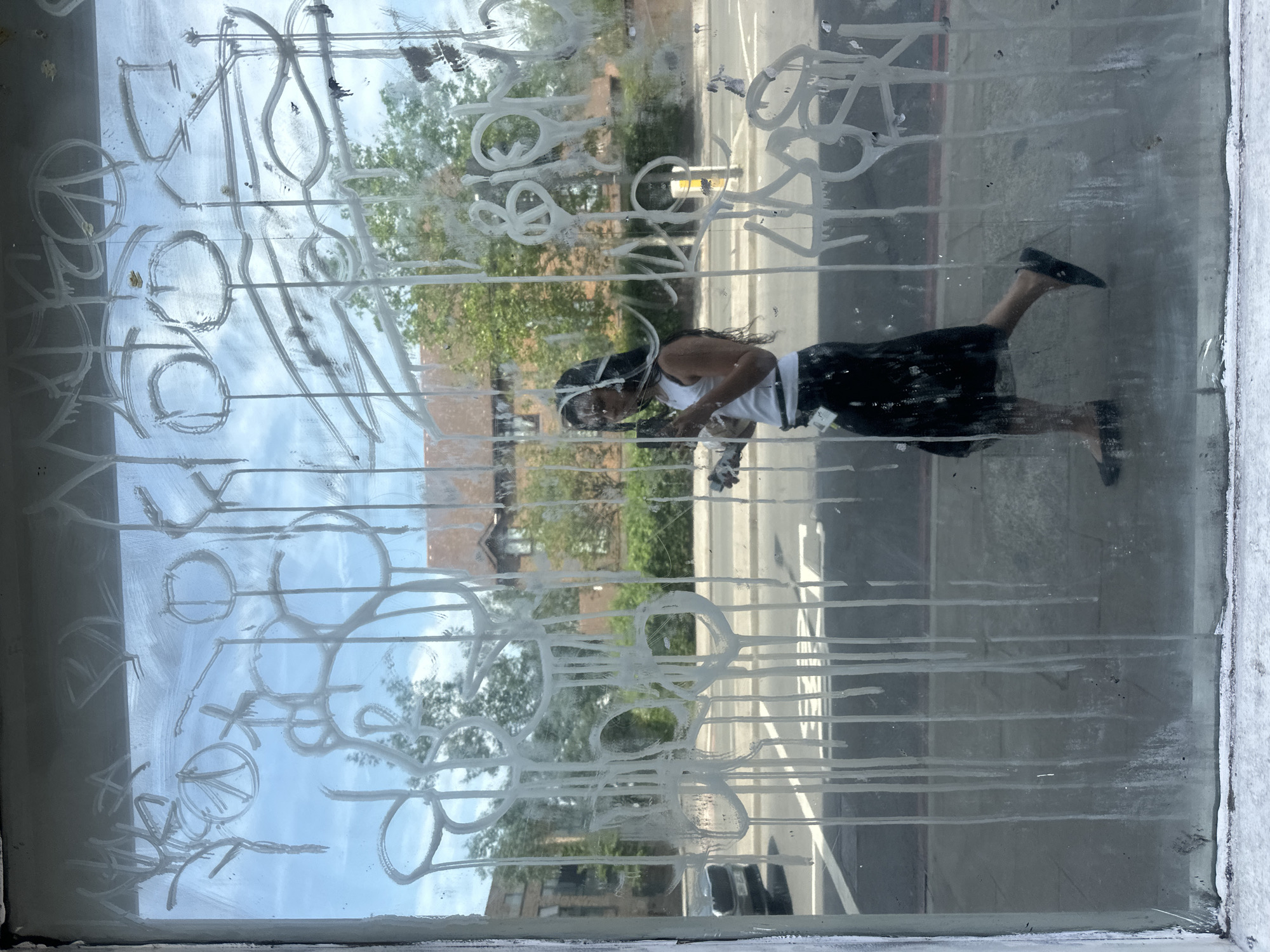 A medium, eye-level shot shows a person cleaning a window. The window is covered with white, graffiti-like markings. A street and buildings are visible through the window. The person, likely in their 20s or 30s, wears a white tank top, a dark skirt, and dark shoes. They have long, dark hair. They are facing left, scrubbing at the window with a cleaning tool in both hands. The setting appears to be an urban environment. Based on the style of buildings and roads visible through the window, the location is possibly in London, United Kingdom.