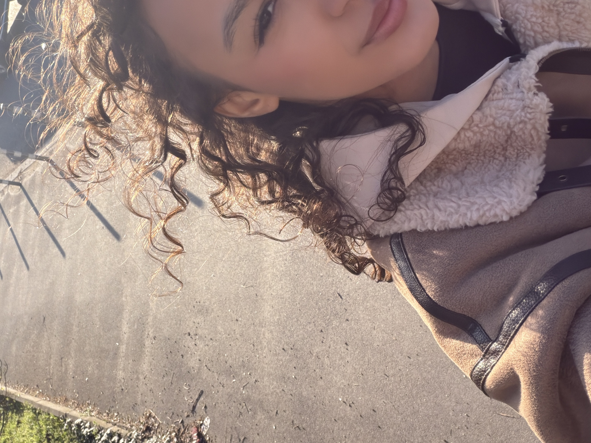 A female individual is partially depicted in a self-portrait, facing the camera on a paved path in Sofia, Bulgaria. The subject's upper right torso and the right side of her face are visible under bright natural light. Her dark, curly hair, highlighted with reddish-brown tones, appears wind-swept. Her right eye is open, enhanced with eyeliner and long lashes. A subtle pink blush is visible on her right cheek, and her lips are slightly parted in a gentle smile, displaying a pink hue. A gold-colored hoop earring is present in her right ear. She wears a light brown or beige jacket with dark faux-leather trim on the shoulder and a cream-colored faux-shearling collar and inner lining, over a dark garment. The background consists of a grey asphalt or concrete path, bordered on the left by a curb and a strip of green grass with dry leaf debris. Long, thin shadows are cast across the upper right portion of the path, indicative of a sunny outdoor environment.