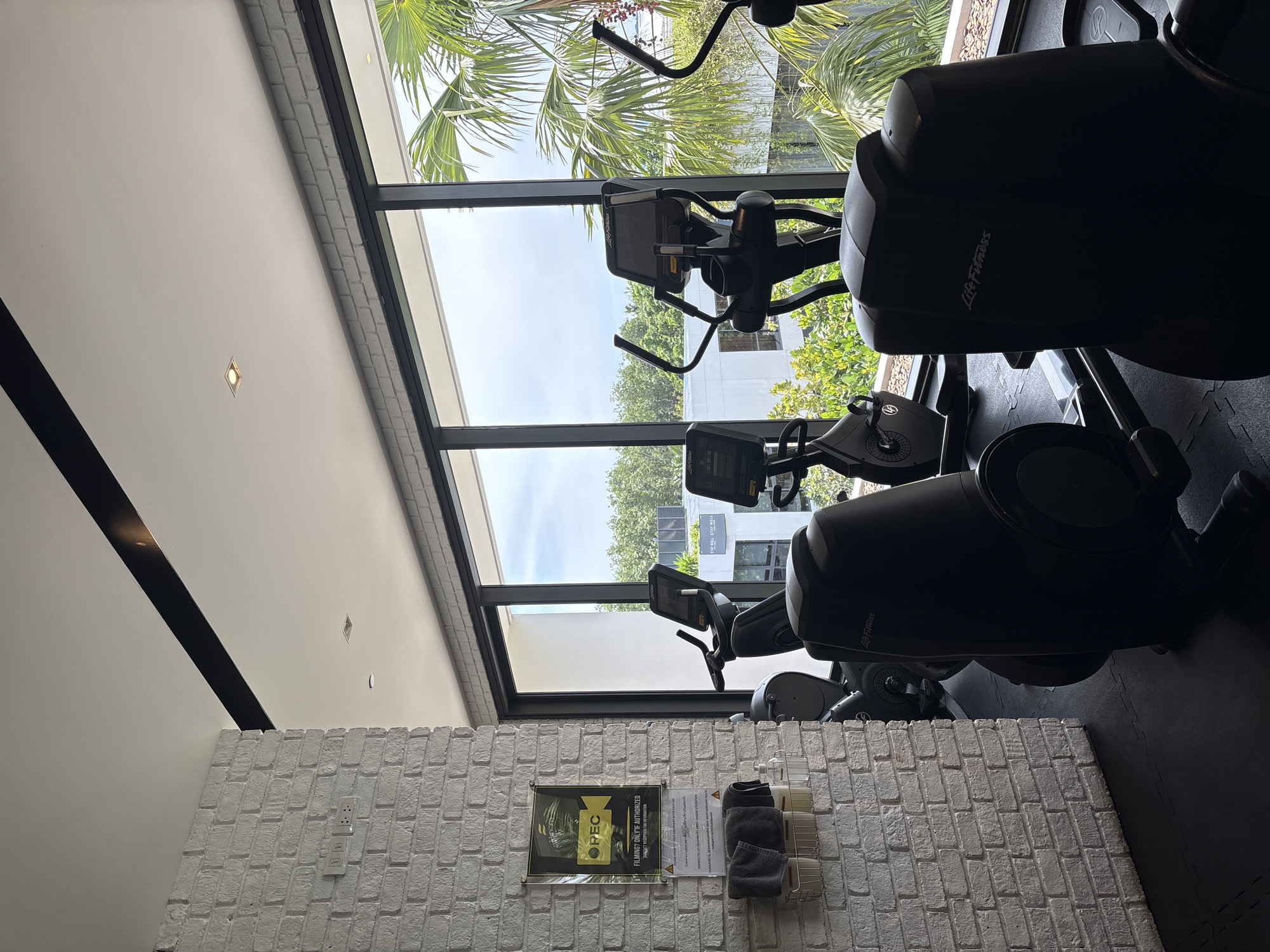 The image depicts the interior of a modern gym in Rawai, Thailand. The gym features several cardio machines, including ellipticals and stationary bikes, positioned in front of a large window offering a view of lush tropical foliage and modern architecture. The walls are a combination of white painted brick and smooth white surfaces. A black beam runs across the ceiling, and recessed lighting is visible. On the left, a white brick pillar displays a sign with a camera icon and text stating "FILMING? ONLY IF AUTHORIZED SUBJECT RECEPTION FOR THE OPERATION." Below the sign, there is a wire basket holding rolled-up towels and a pump bottle, likely hand sanitizer or soap. The floor is covered with black interlocking mats. The overall atmosphere is clean and contemporary, with natural light filtering in from the large windows. No people are present in the image.
