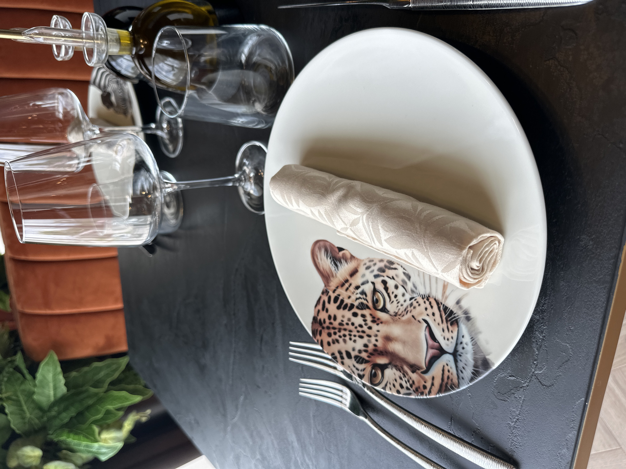 The scene is a restaurant table set for a meal. There are three wine glasses and a bottle of wine. A white plate decorated with a leopard's head has a rolled-up napkin on it. A fork and knife are placed next to the plate. A small potted plant is visible in the lower left corner. The table is black and located indoors, in Sofia, Bulgaria.